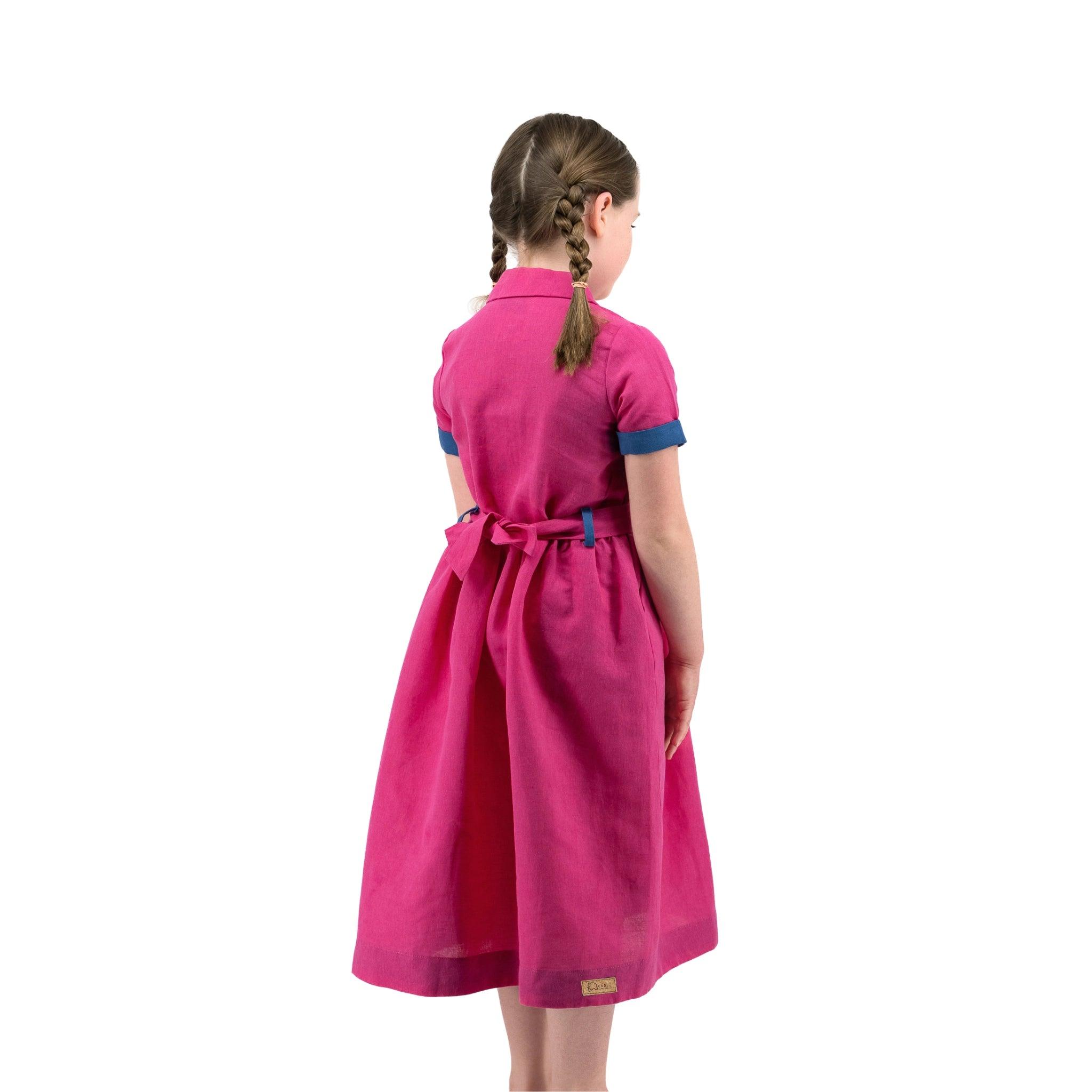 A young girl in a Karee fuchsia purple linen dress with a bow tied at the back, standing in a side pose, looking to her left, isolated on a white background.