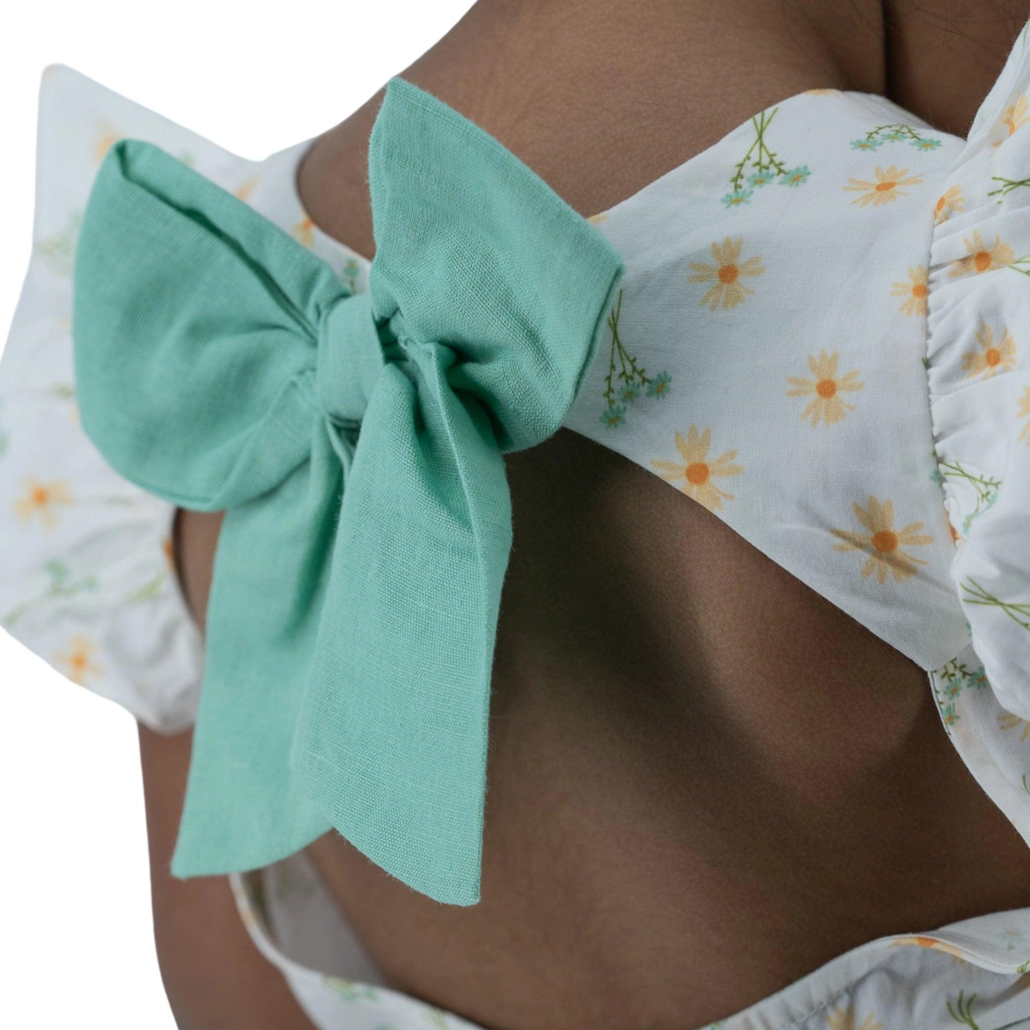 Close-up of a person wearing a Karee Petite Blossom Cotton Dress in Smoked Pearl with yellow floral print and a large green bow on the back.