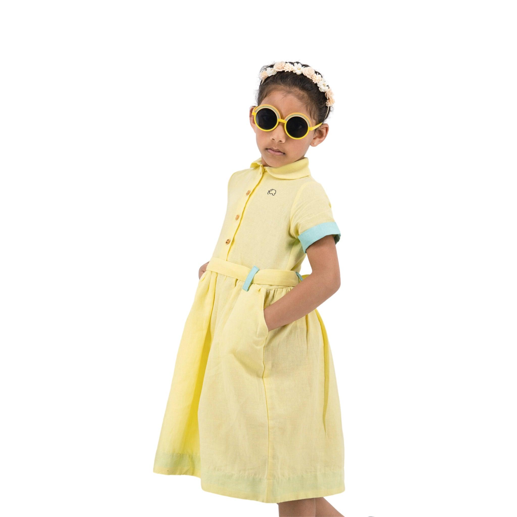 Young girl posing in a stylish Karee elfin yellow linen below knee length dress and sunglasses, with a white floral headband, against a white background.