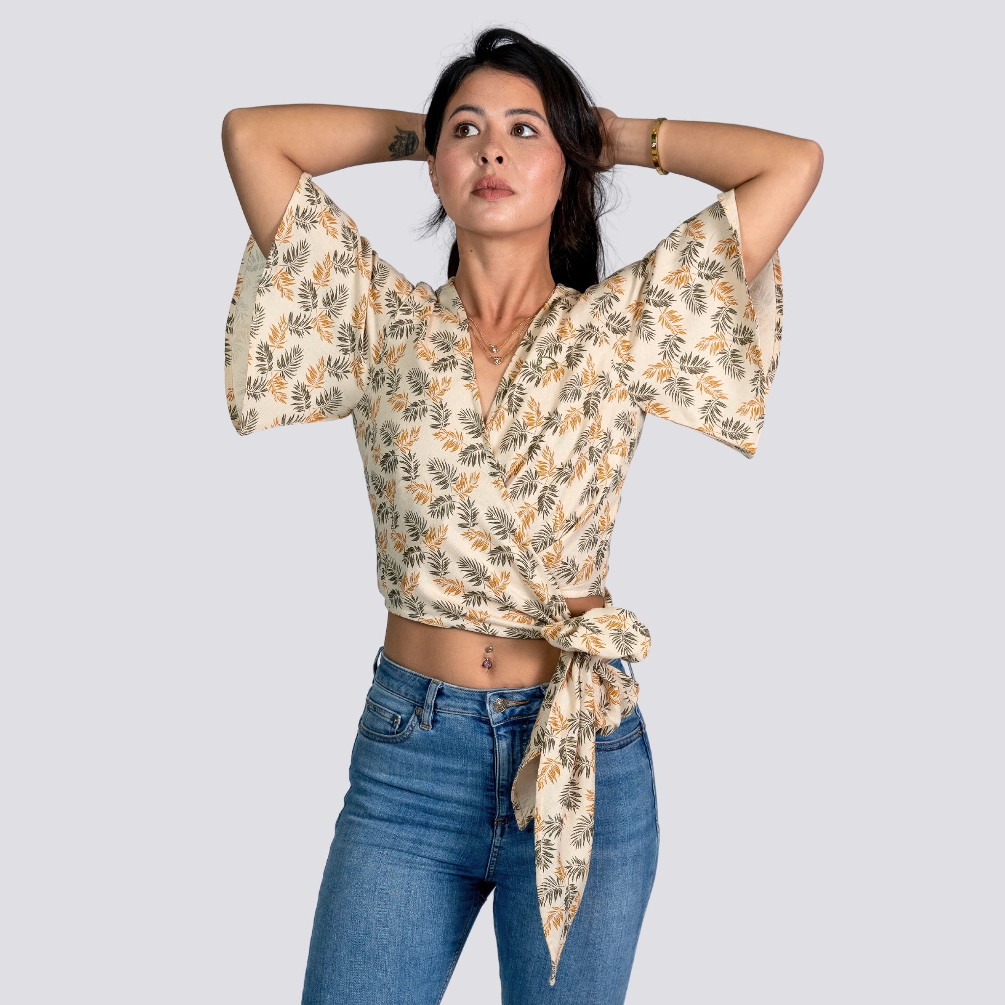 Woman in a ChicPalms Women's Eco-Friendly Wrap Top - Biscuit Bliss by Karee and jeans looking thoughtfully upwards, hands behind her head, against a neutral background.