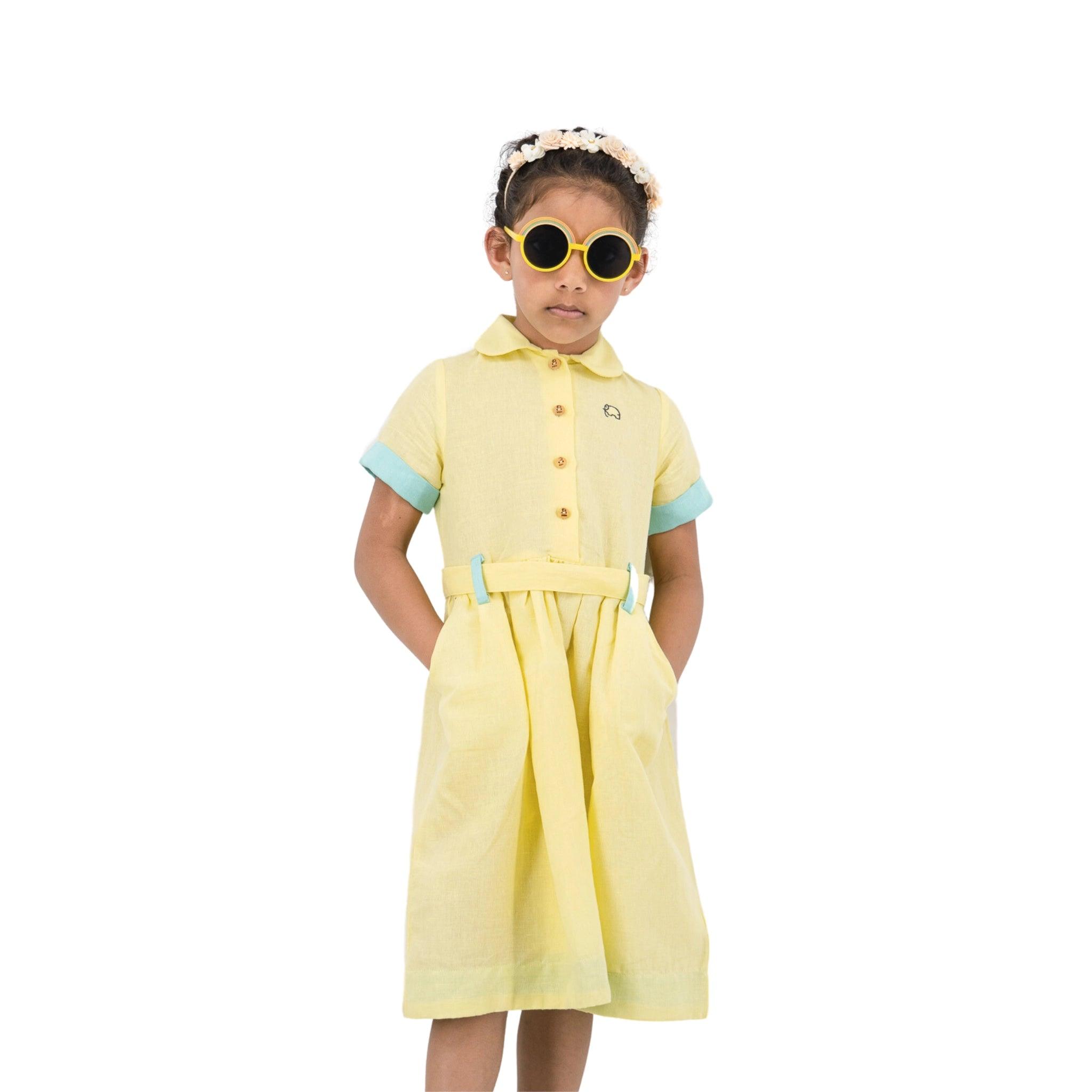 Young girl wearing a Karee elfin yellow linen below knee length dress for girls and sunglasses, standing confidently with her hands on her hips, isolated on a white background.