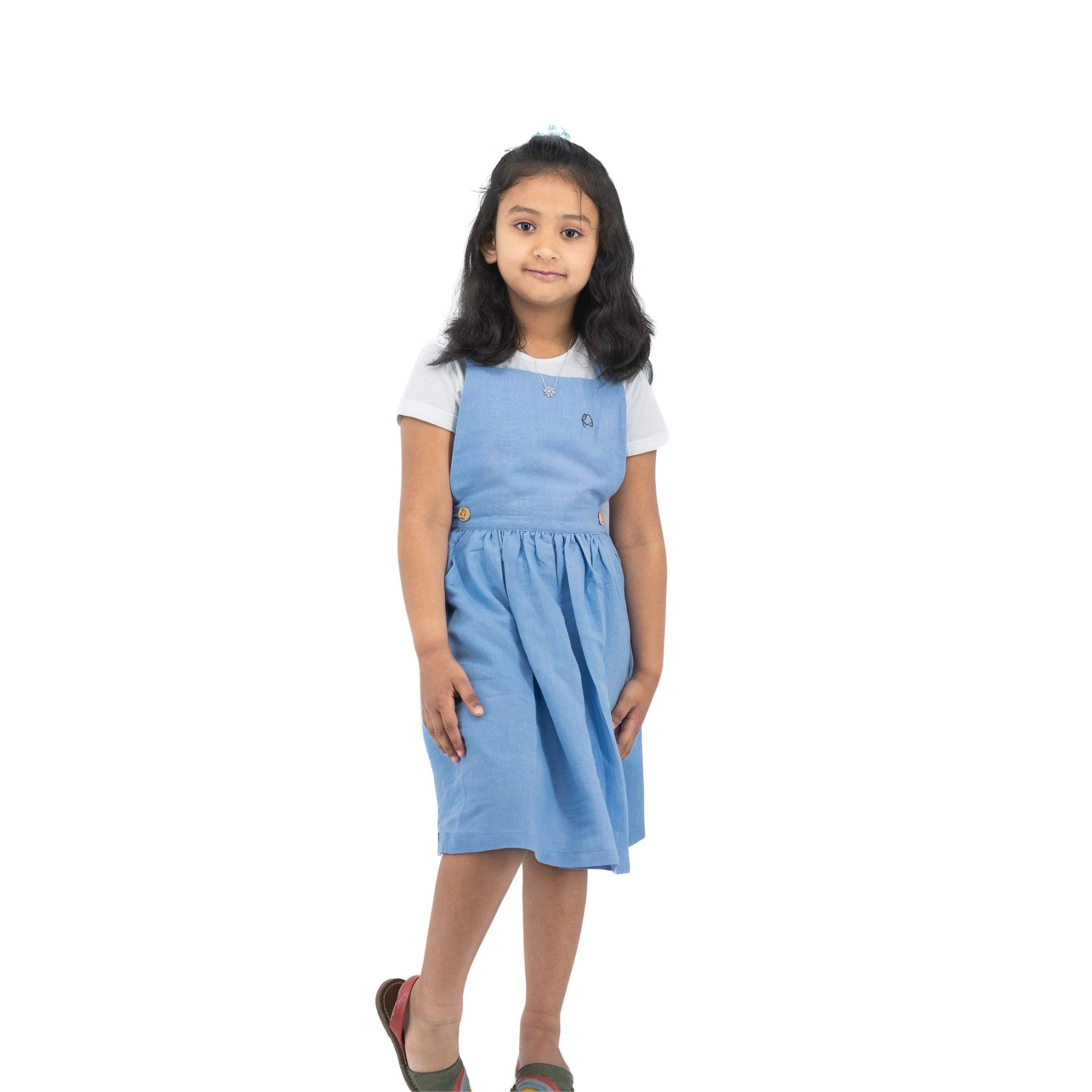 Young girl in a Karee cerulean blue linen pinafore standing alone against a white background, looking at the camera with a slight smile.