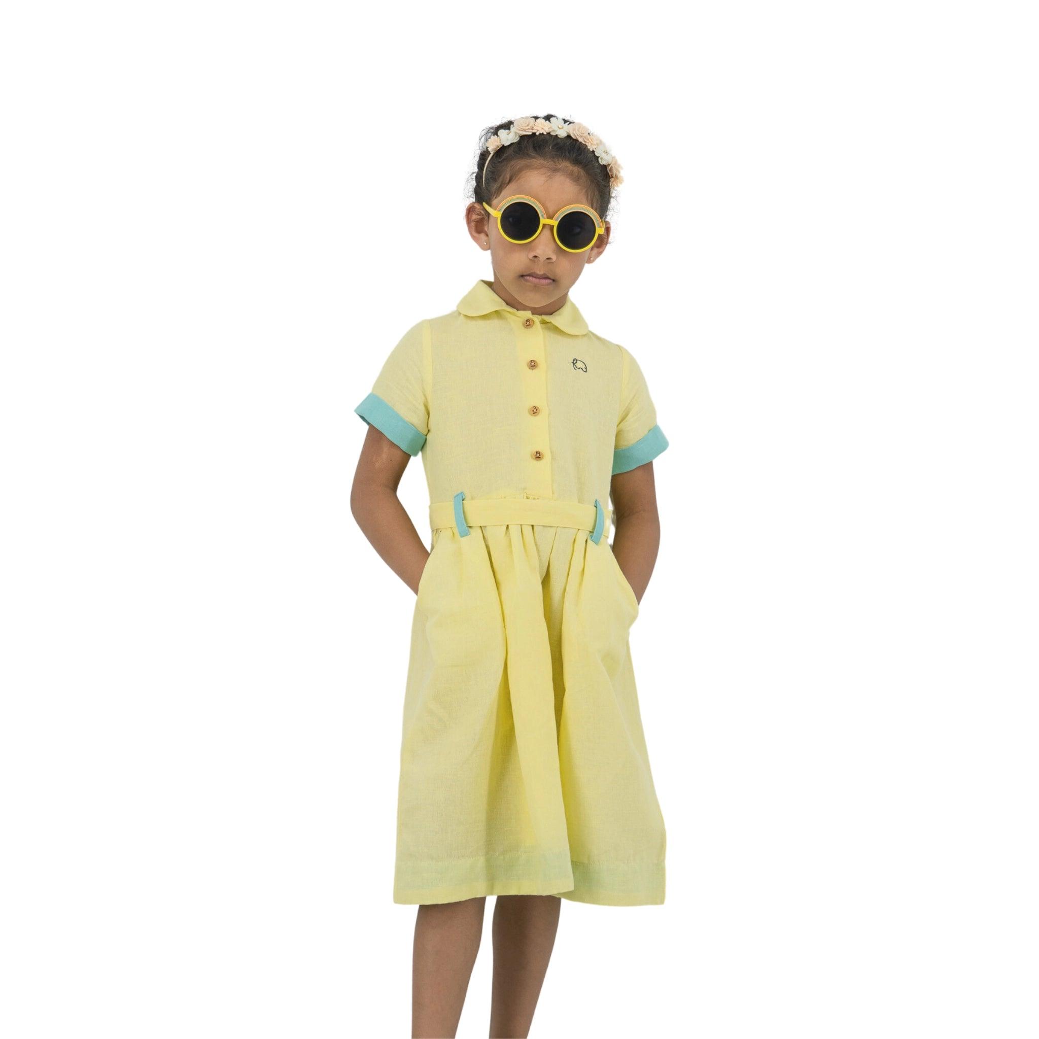 Young girl in a Karee Elfin Yellow Linen Below Knee Length Dress for Girls with blue trim, wearing yellow sunglasses and a white floral headband, standing against a white background.