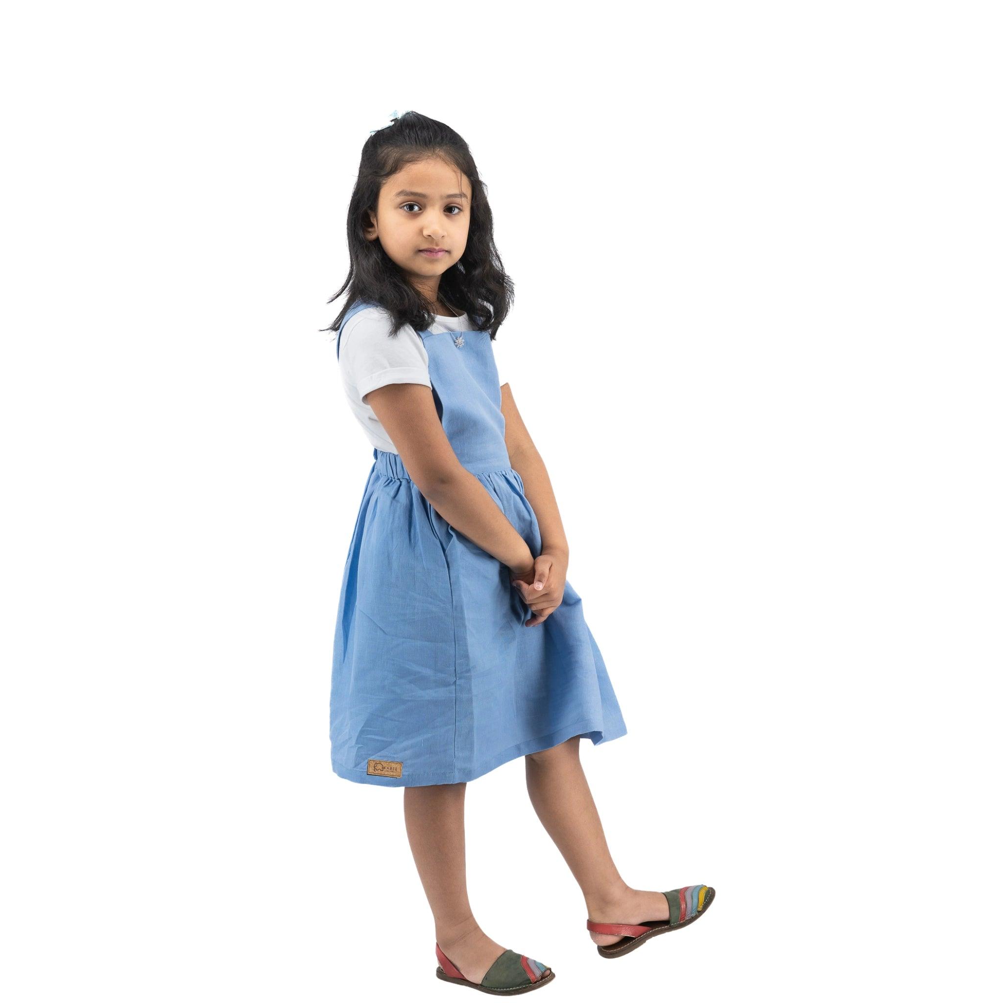 Young girl in a Karee cerulean blue linen pinafore and colorful shoes standing sideways and looking at the camera, isolated on a white background.