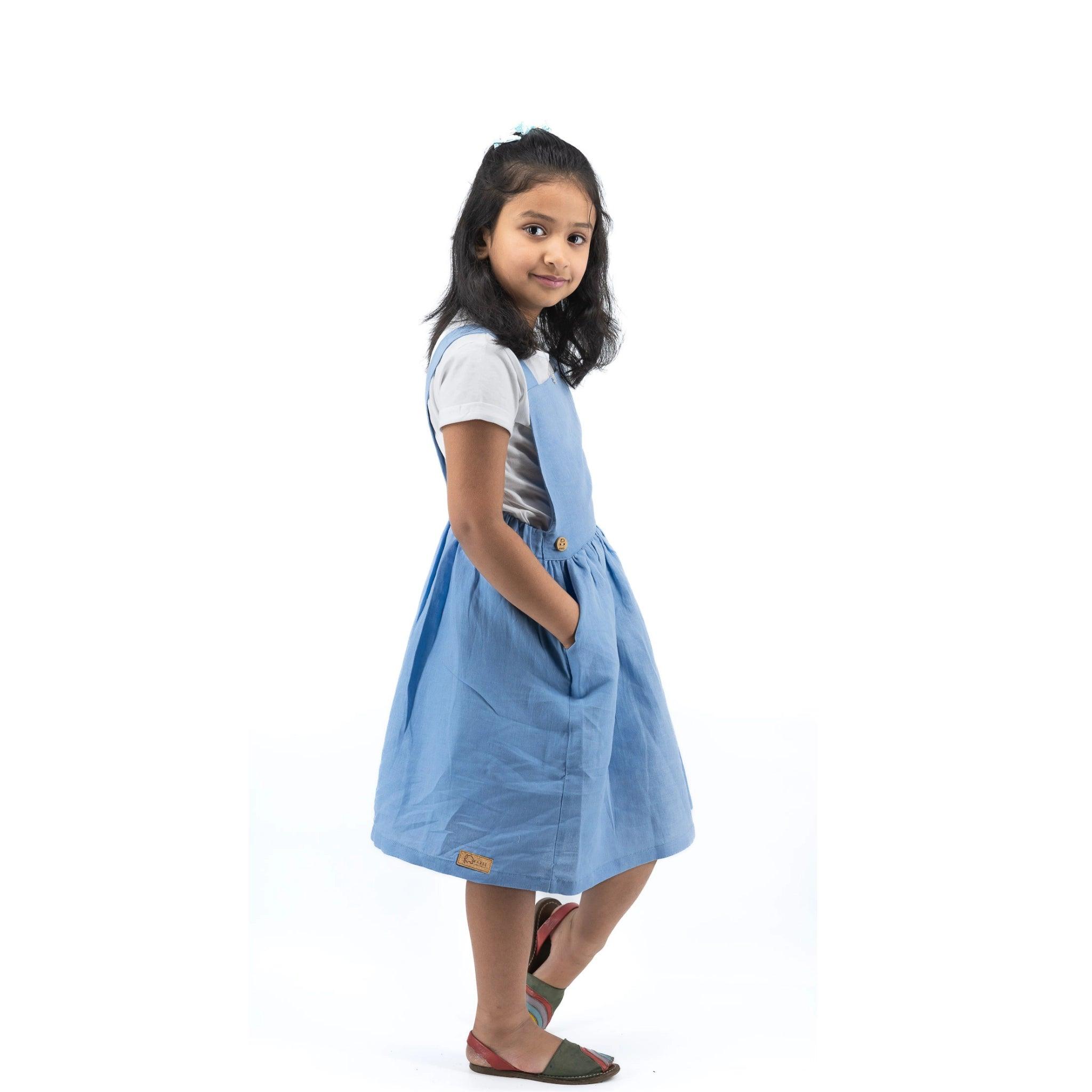 A young girl in a Karee cerulean blue linen pinafore and white shirt posing with her hand on her hip, looking over her shoulder on a white background.