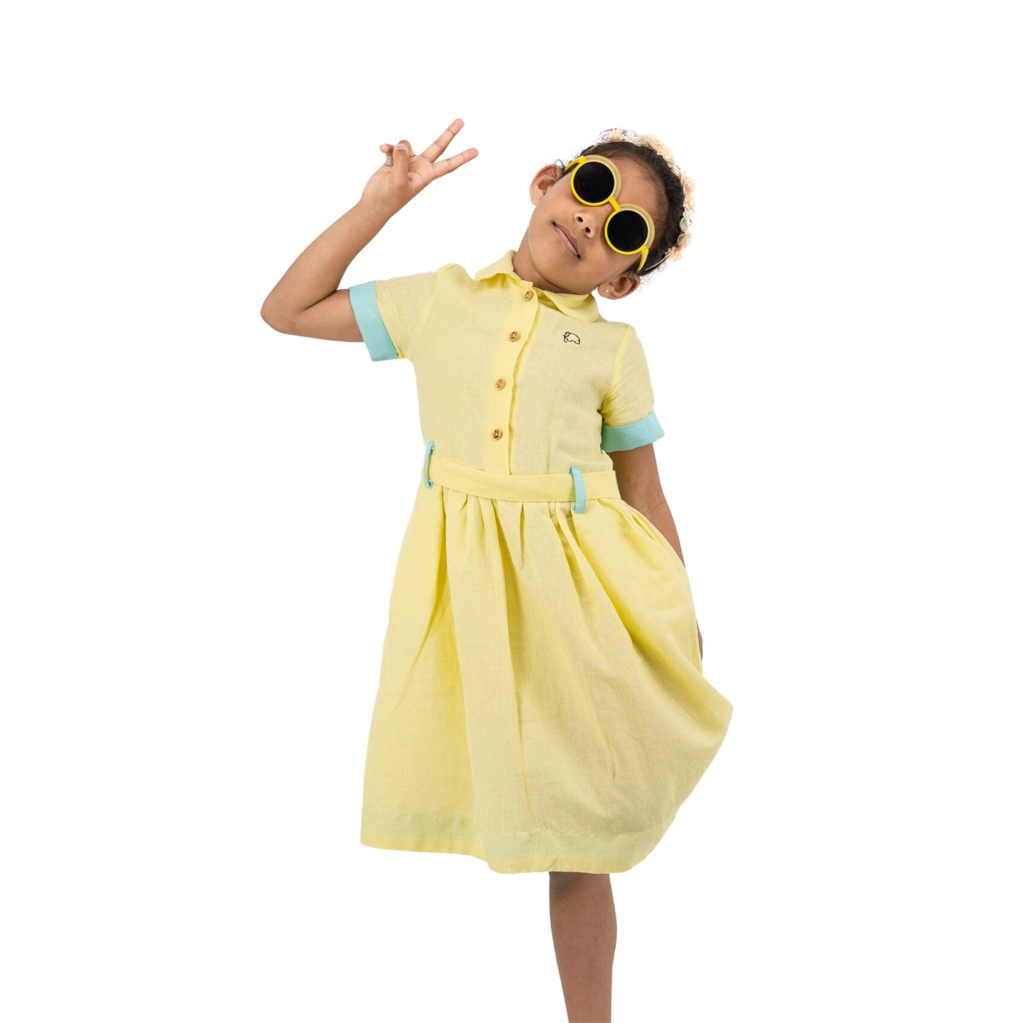 A young girl in a Karee Elfin Yellow Linen Below Knee Length Dress for Girls and sunglasses gives a peace sign, standing against a white background.