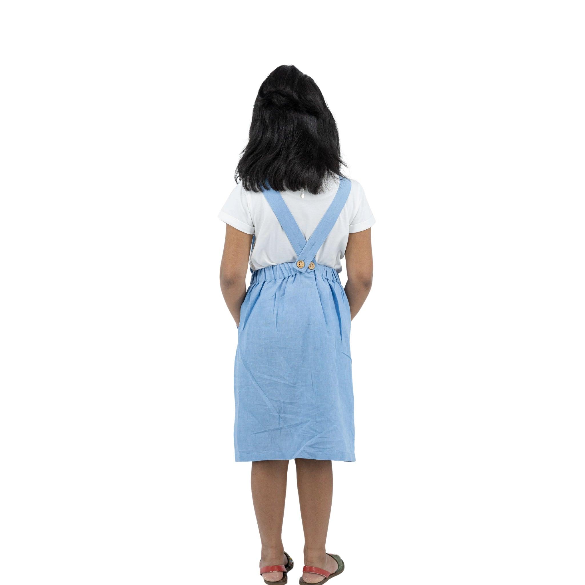 A woman stands with her back to the camera, wearing a white t-shirt and a Karee cerulean blue linen pinafore, isolated on a white background.