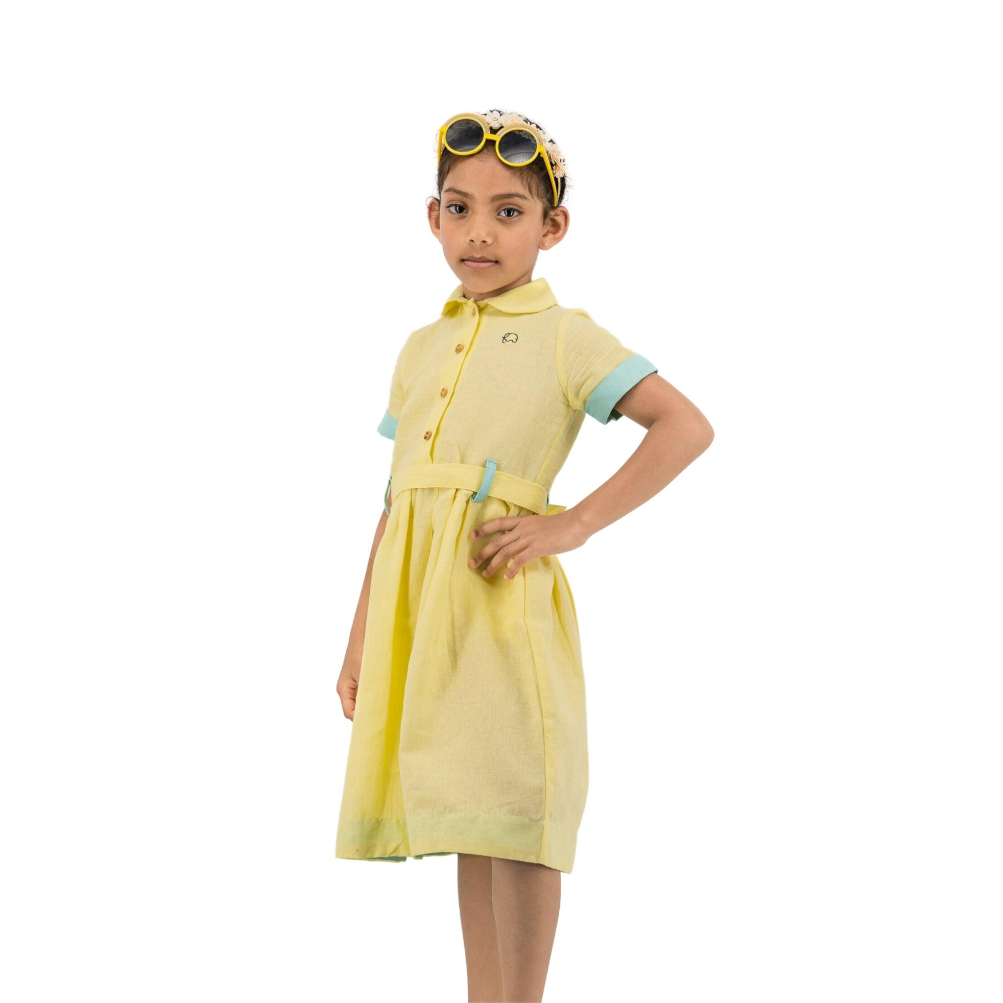 A young girl stands confidently, wearing a Karee elfin yellow linen dress and blue-trimmed goggles on her head, isolated on a white background.
