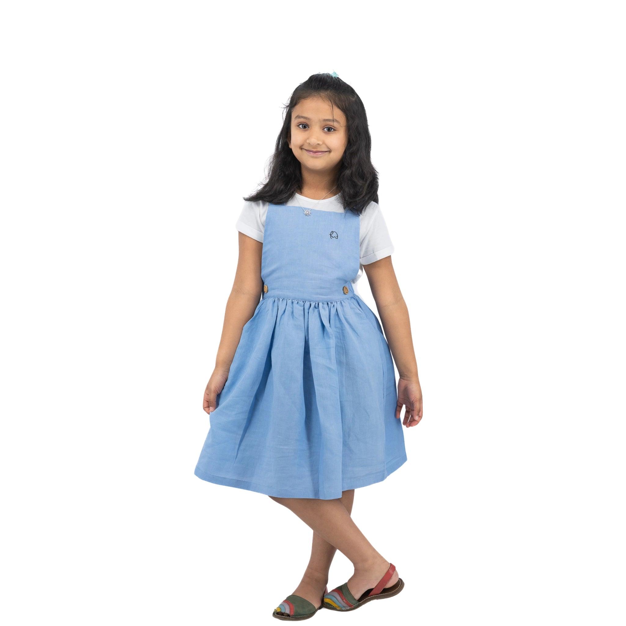 A young girl in a Karee cerulean blue linen pinafore standing and smiling against a white background.