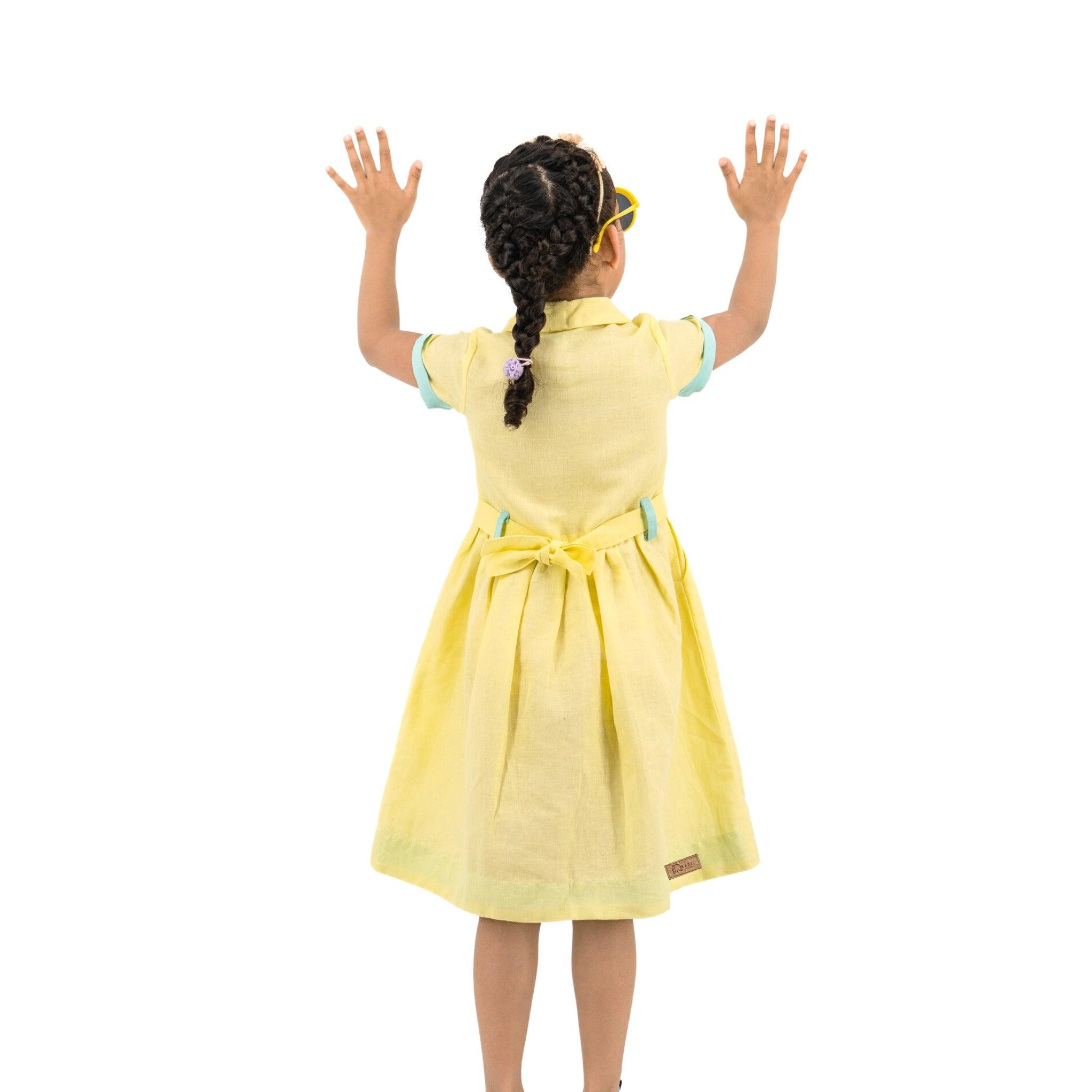 Back view of a young girl with braided hair, wearing a Karee Elfin Yellow Linen Below Knee Length Dress for Girls and yellow glasses, standing with her hands raised against a white background.