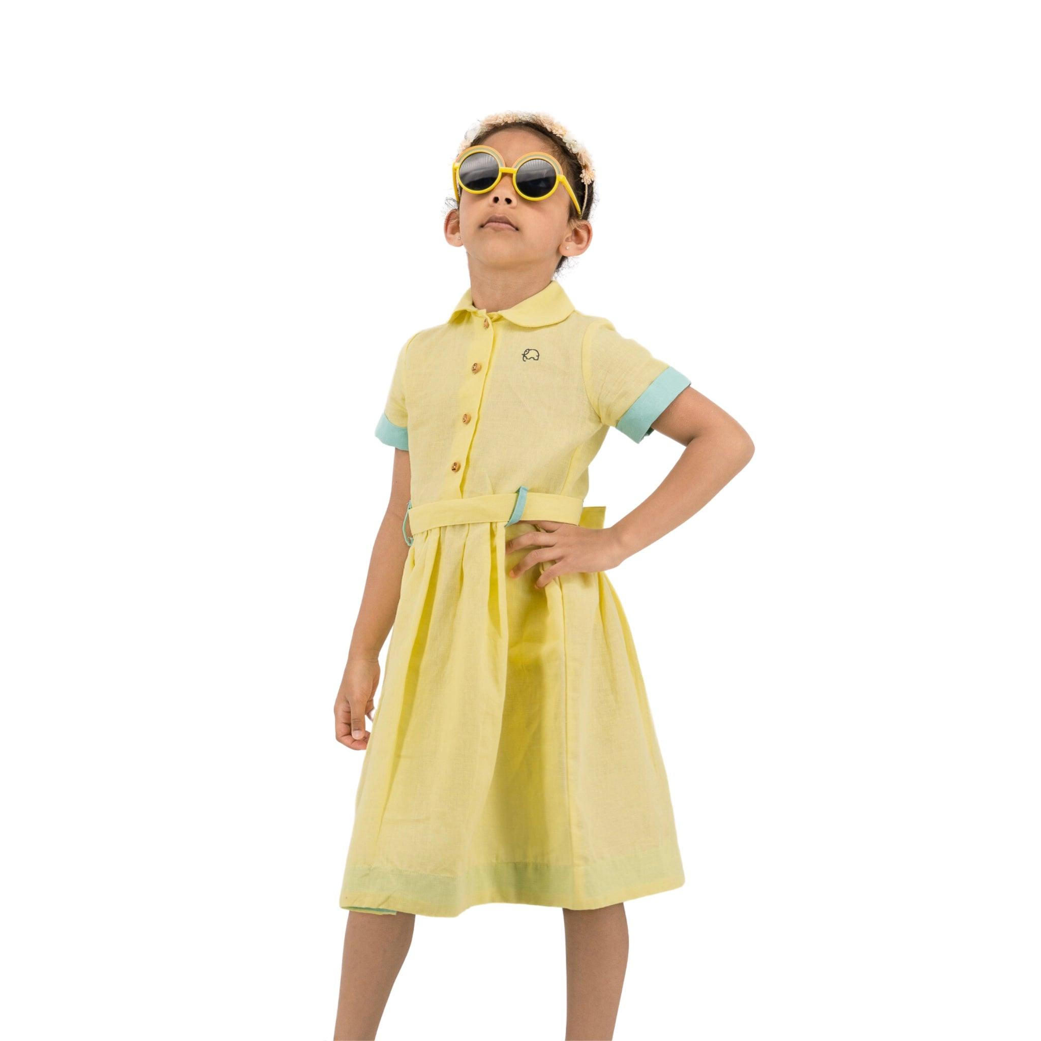Young girl wearing a Karee elfin yellow linen below knee length dress and large sunglasses, hands on hips, posing confidently against a white background.