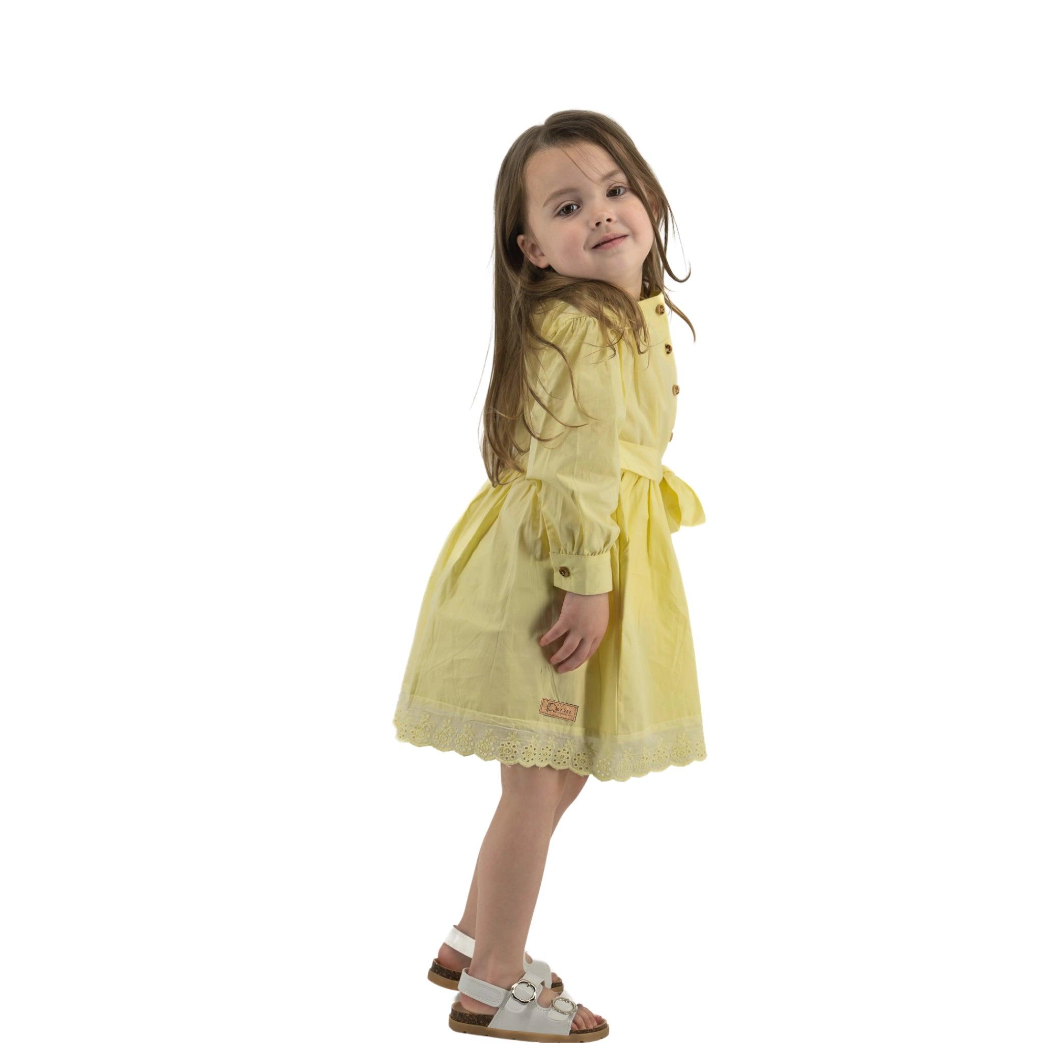 Young girl in a Karee yellow long puff sleeve cotton dress looking over her shoulder with a slight smile, isolated on a white background.