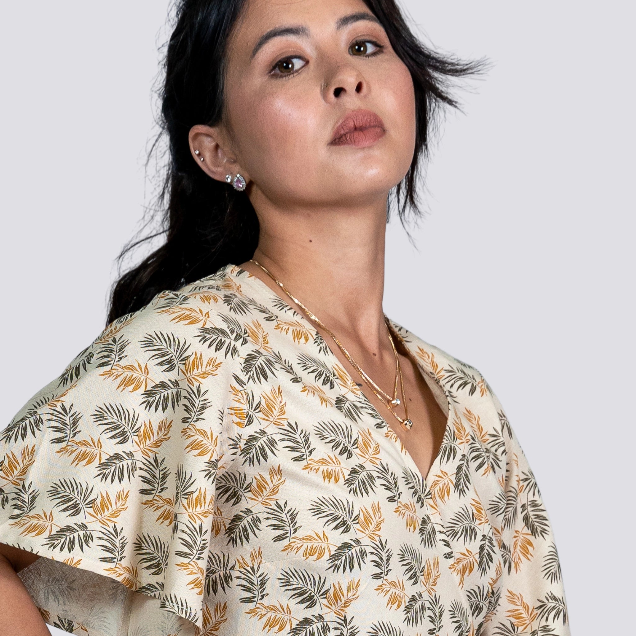A woman with dark hair wearing a ChicPalms Women's Eco-Friendly Wrap Top in Biscuit Bliss by Karee, along with subtle jewelry, looking confidently off-camera against a light grey background.