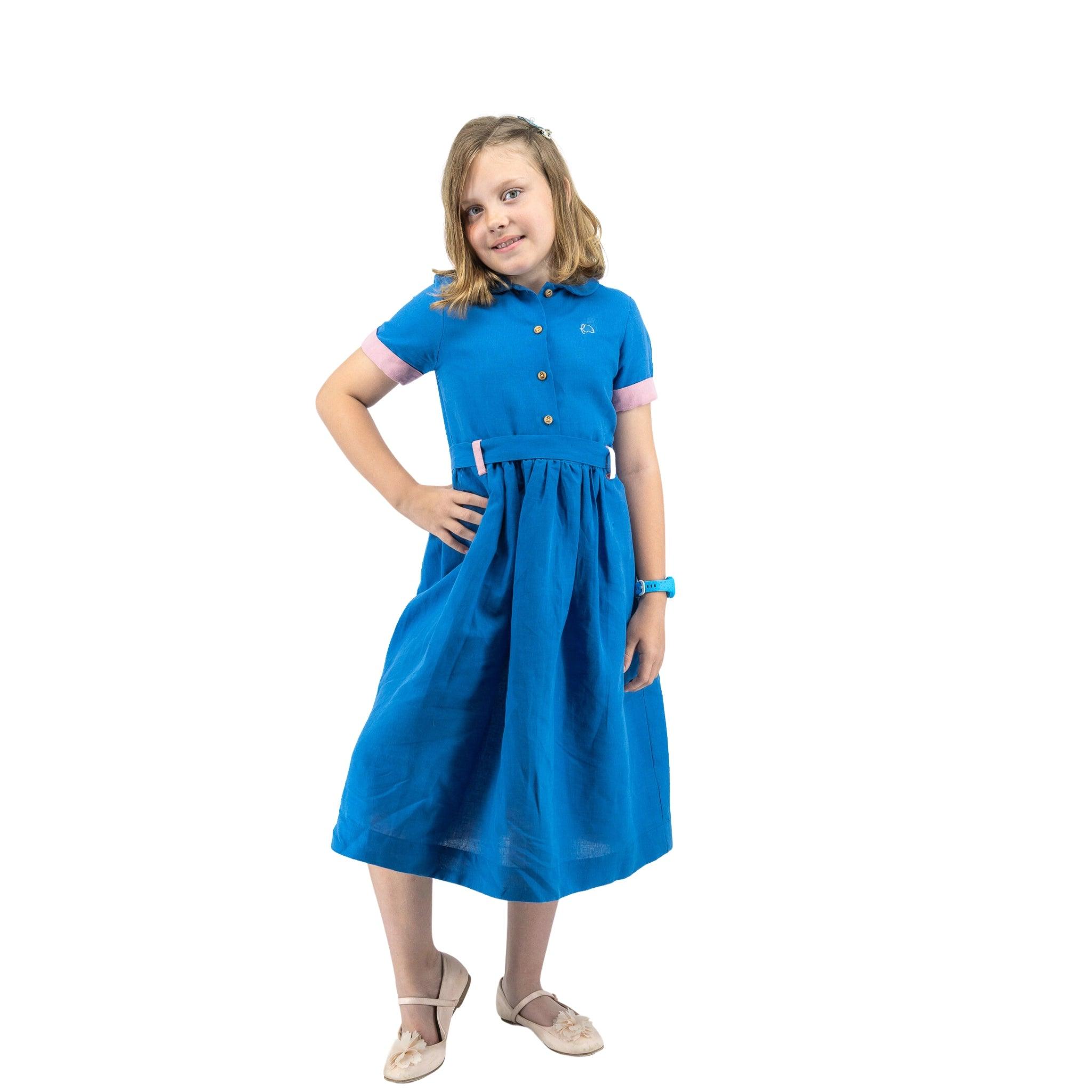 A young girl stands confidently, hands on hips, wearing a Karee Parisian Blue Linen Dress for Girls and beige shoes against a white background.