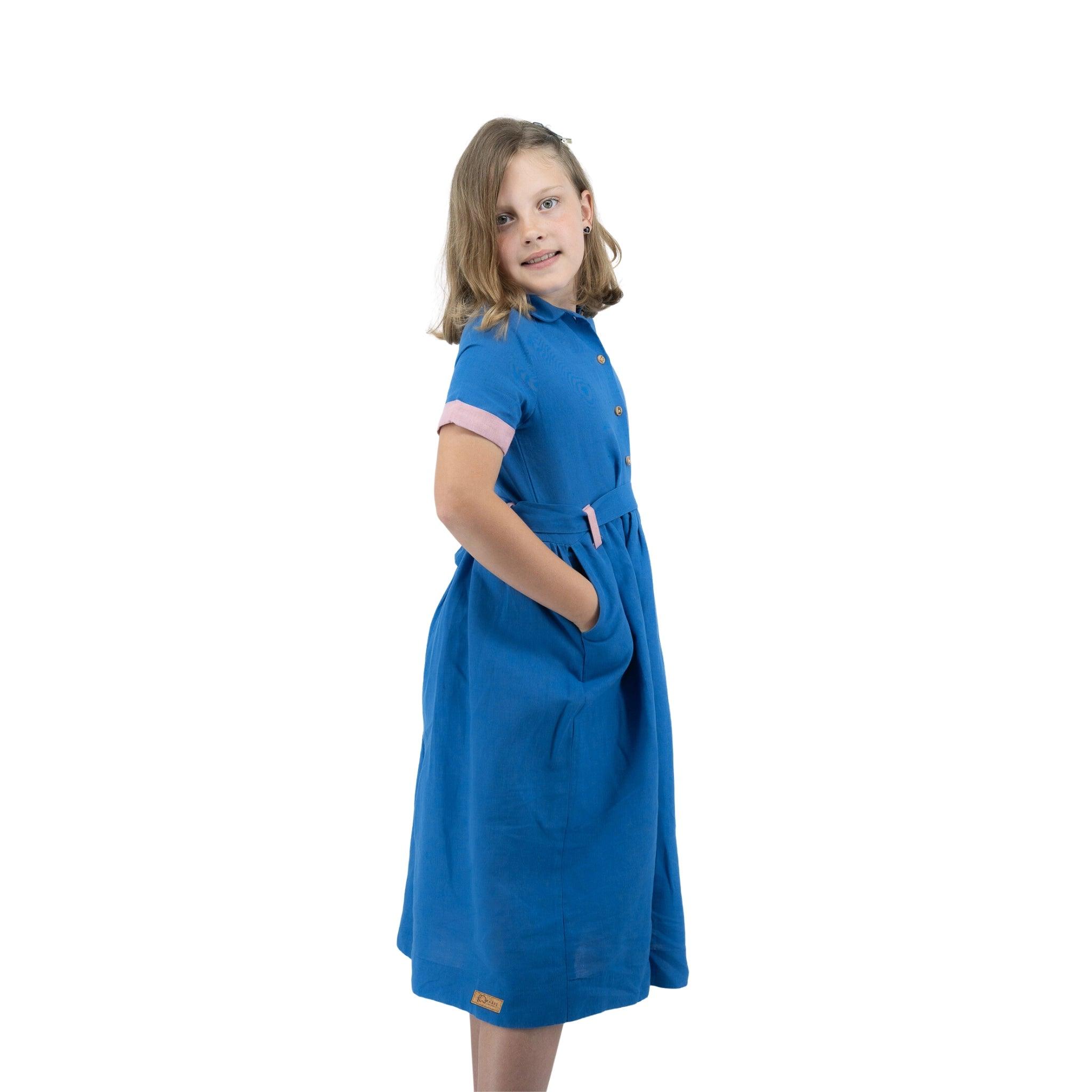 Young girl posing in a Karee Parisian Blue Linen Dress for Girls with hands on her hips, smiling at the camera, isolated on a white background.