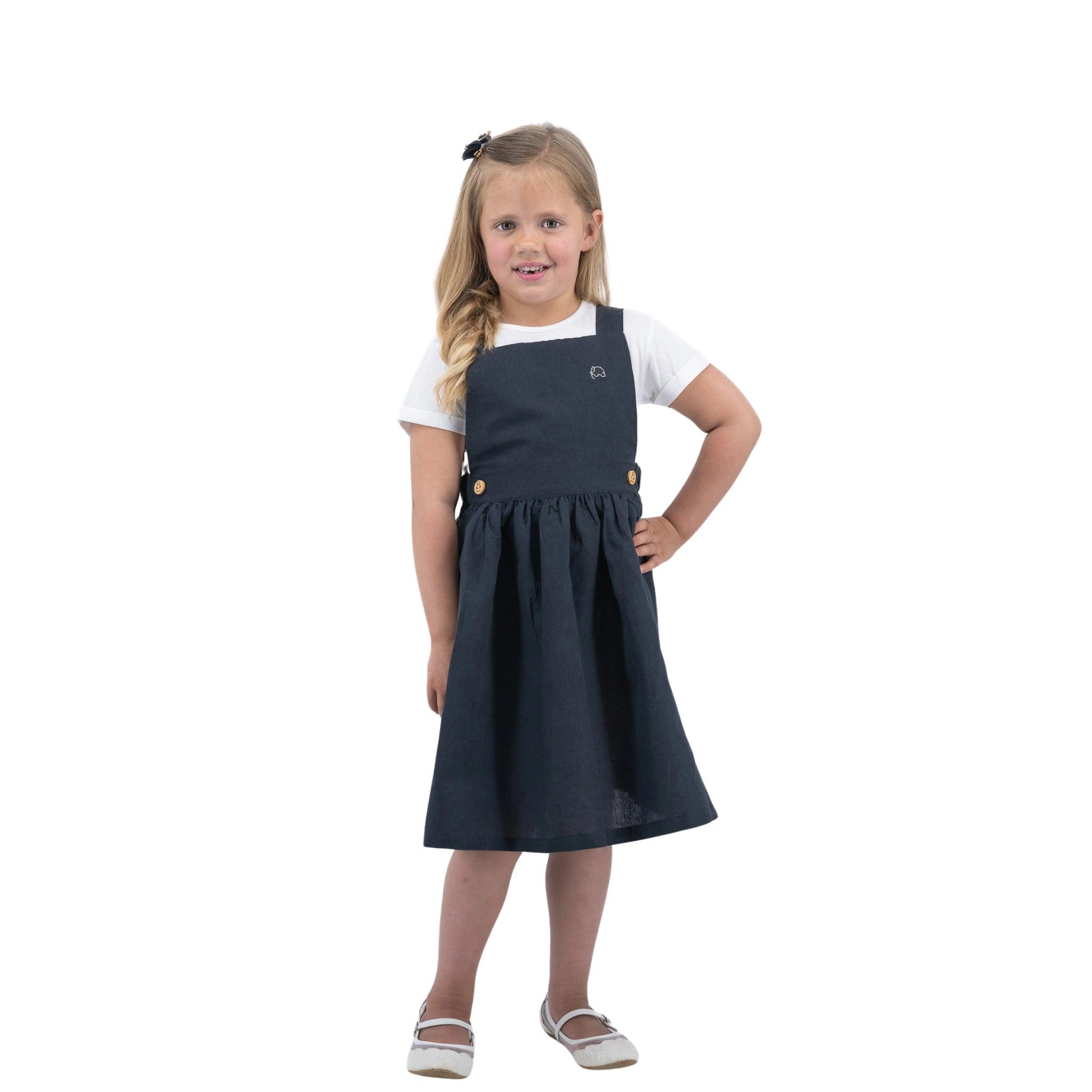 A young girl with blonde hair stands confidently, wearing a Karee Ebony Black Linen Pinafore dress over a white shirt, matched with white shoes.