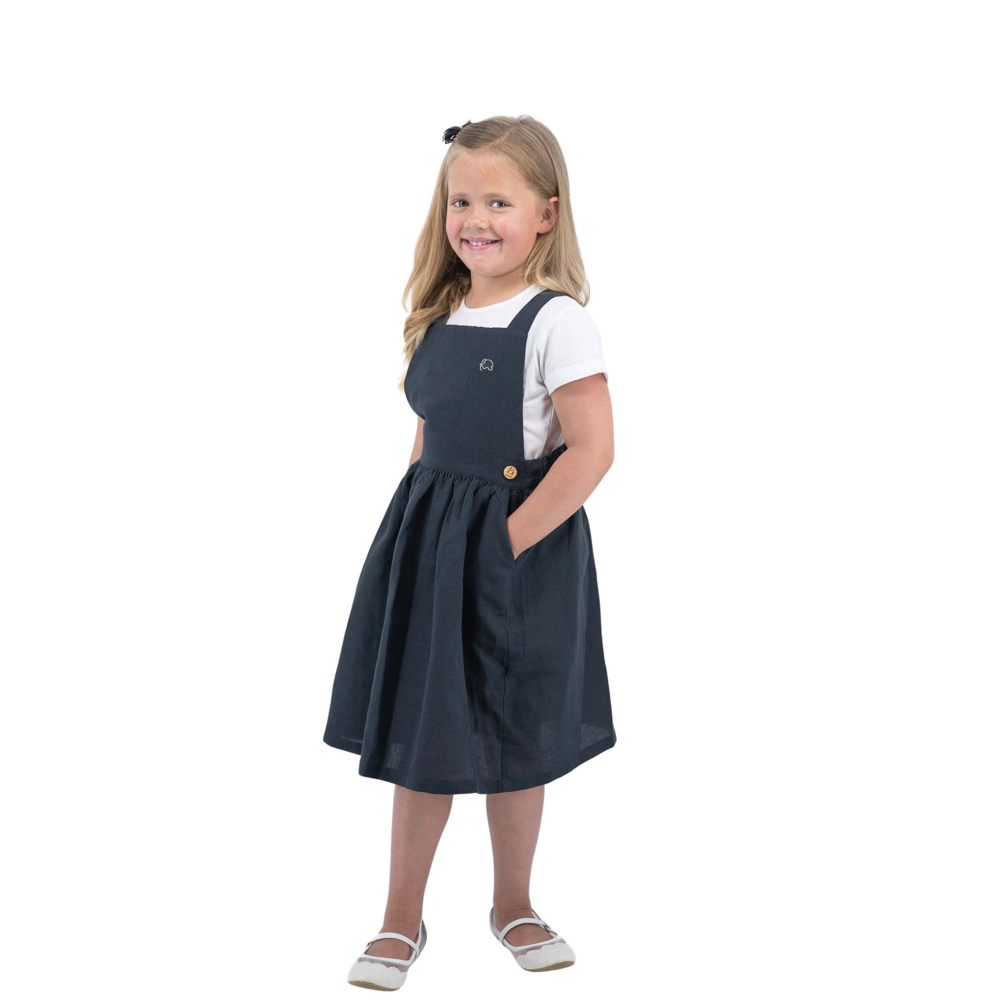 A young girl smiling, wearing a Karee ebony black linen pinafore for girls and white shirt, standing against a white background.