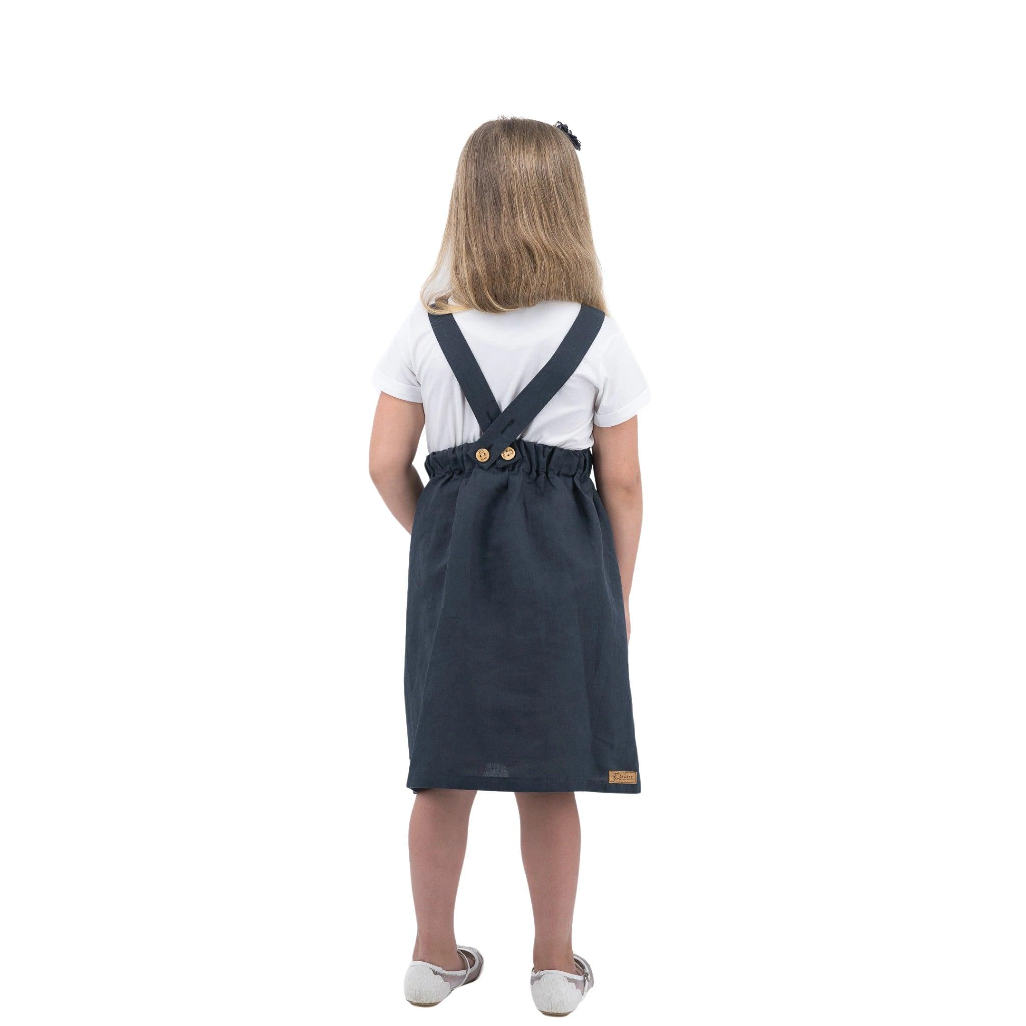 Young girl seen from behind wearing a white t-shirt and a Karee Ebony Black Linen Pinafore for Girls with suspenders, standing against a white background.