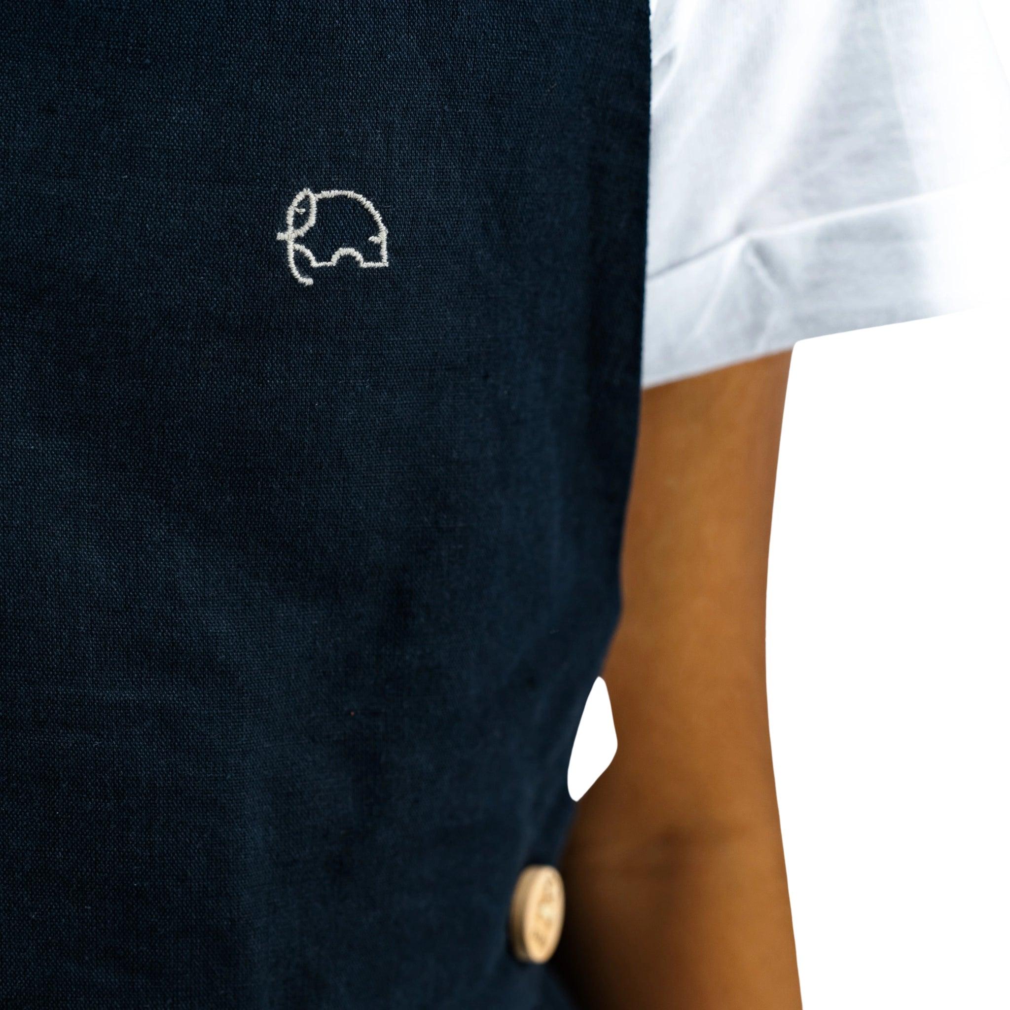 Close-up of a dark blue fabric featuring Karee's Ebony Black Linen Pinafore for Girls with a small white elephant embroidery on the chest of a shirt, partial view of a person's torso.
