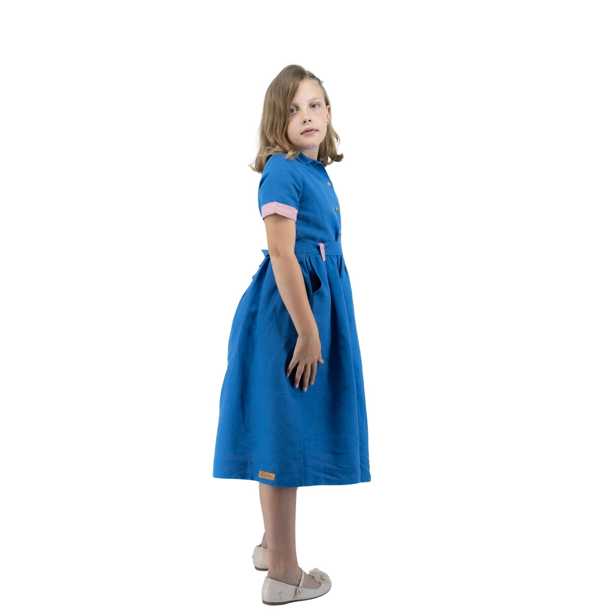 Young girl in a Karee Parisian Blue Linen Dress for Girls and white shoes standing sideways, looking over her shoulder with a neutral expression.