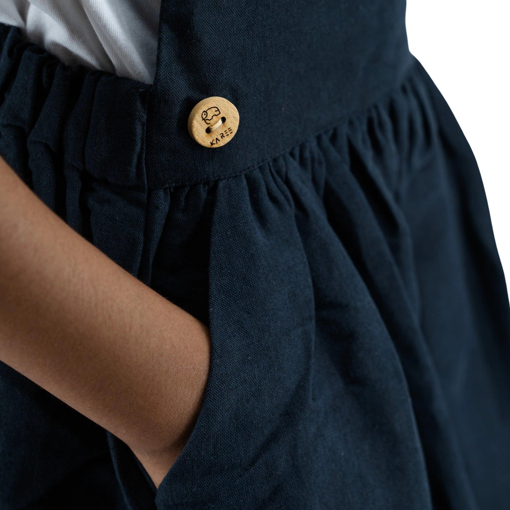 Close-up of a Karee ebony black linen pinafore for girls with a unique button featuring a smiley face design, focused on the waist area where the fabric gathers.