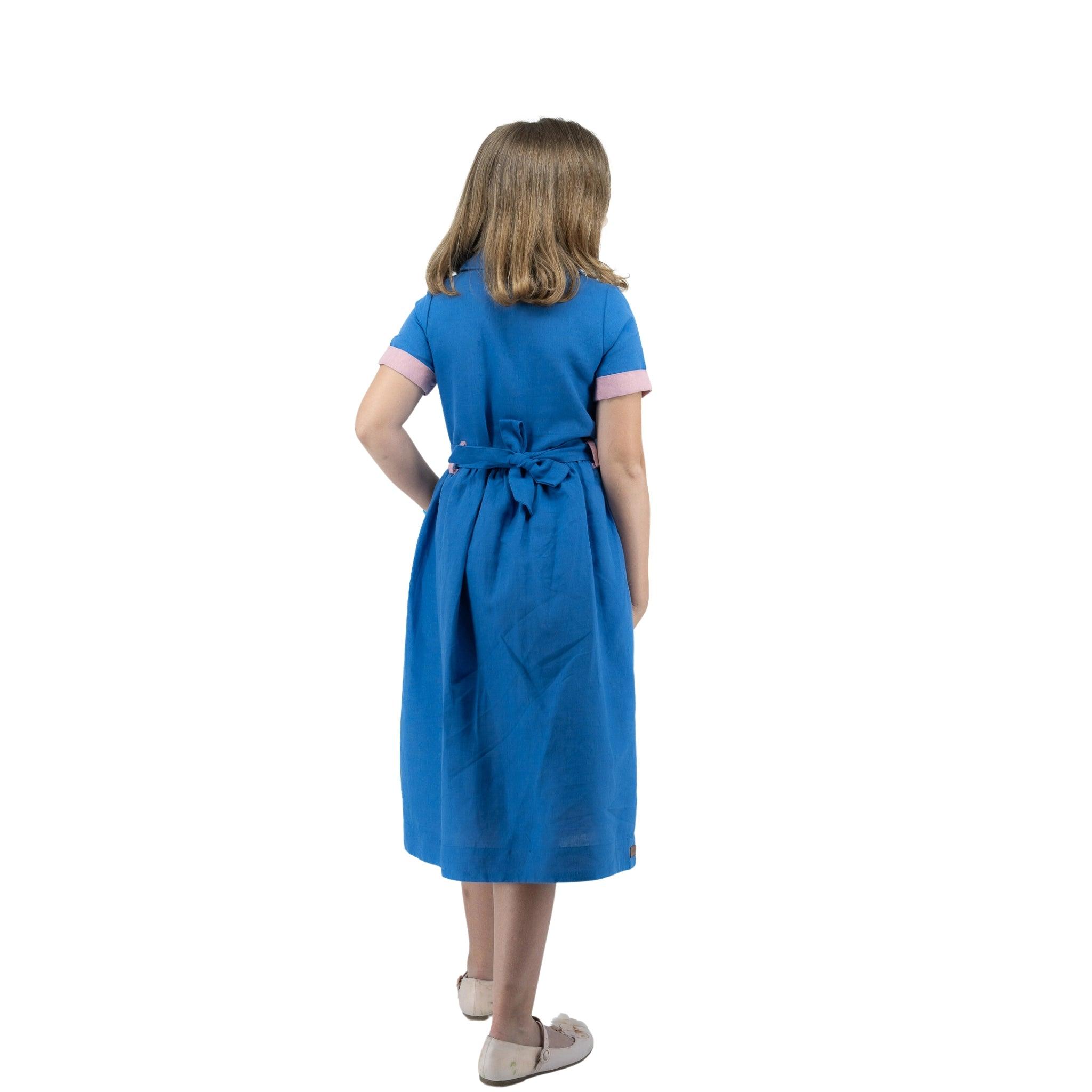 Girl in Karee Parisian Blue Linen Dress for Girls with bow tied at the back, standing facing away from the camera, isolated on white background.