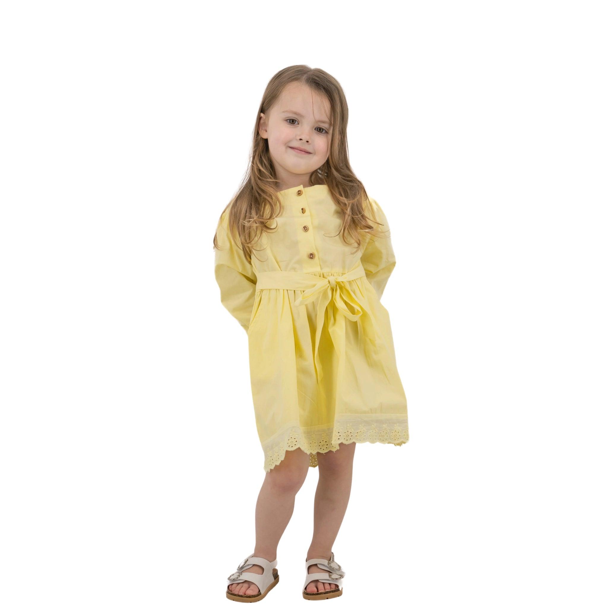 A young girl with long hair in a Karee Yellow Long Puff Sleeve Cotton Dress and sandals, standing and smiling against a white background.