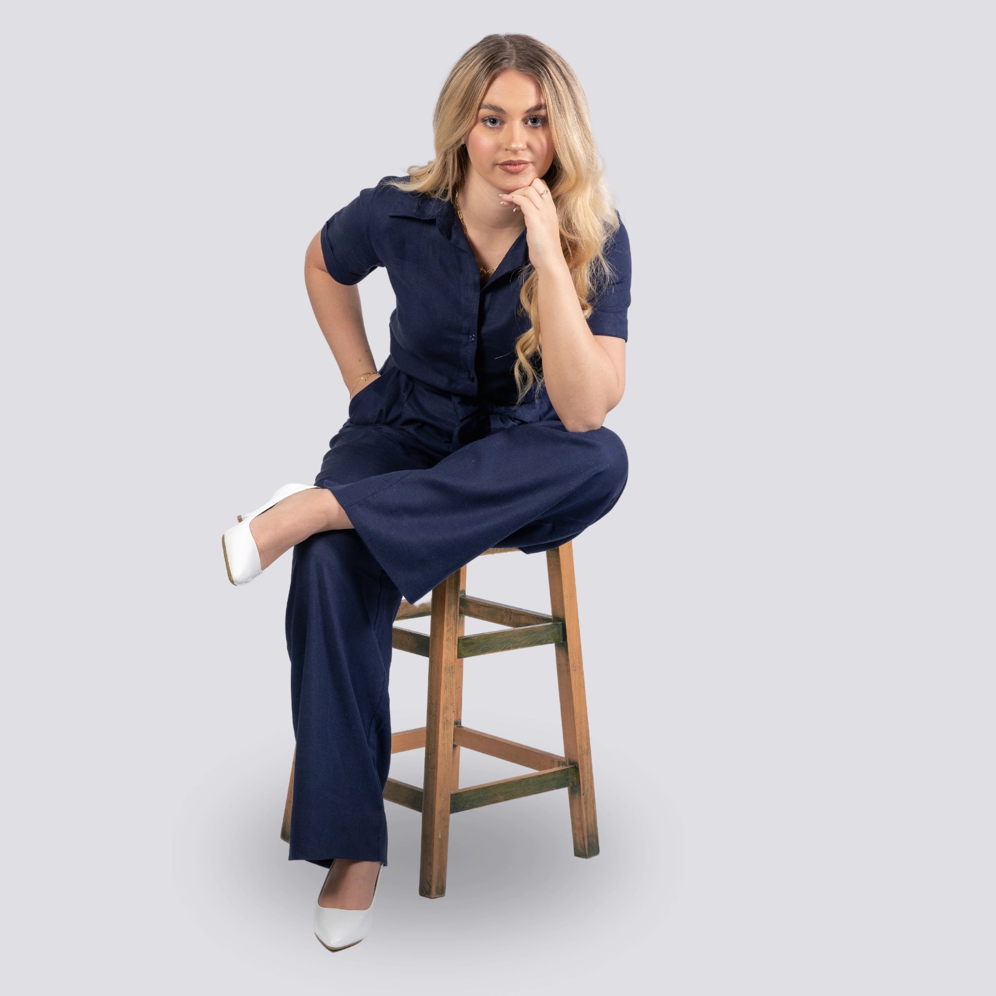 A woman in a Midnight Blue Elegance Viscose Linen Jumpsuit by Karee sitting on a wooden stool against a grey background, looking thoughtful.