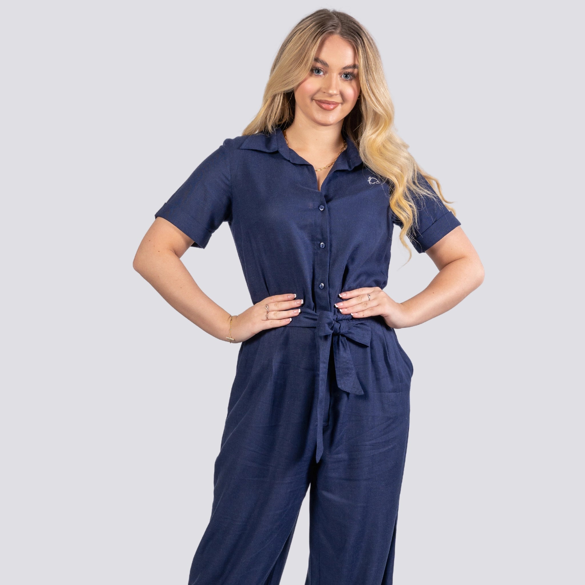 Woman in a Karee Midnight Blue Elegance Viscose Linen Jumpsuit with short sleeves and a tied waist, standing with hands on hips, smiling at the camera against a light gray background.