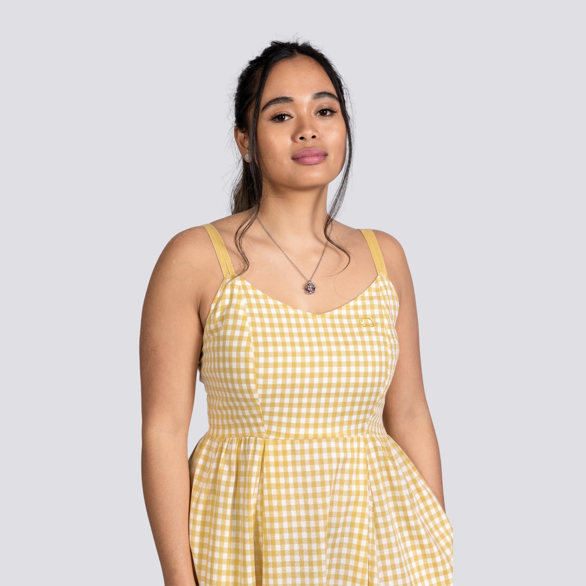 A young woman in a Sunshine Chic Yellow Plaid Cotton Mini Dress by Karee stands against a gray background, looking slightly to her left with a neutral expression.