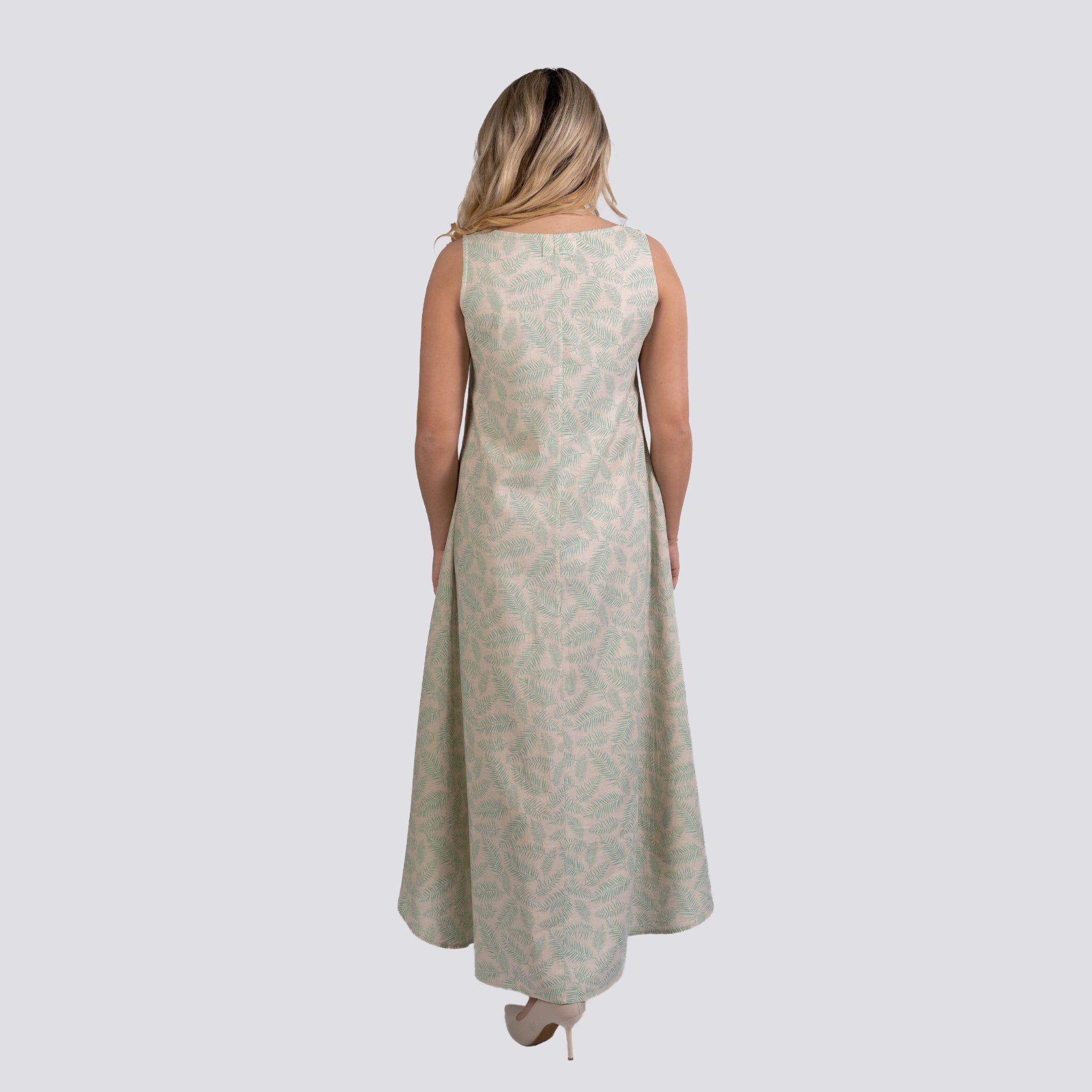 A woman stands with her back to the camera, wearing a long, sleeveless U-neck green dress with a leaf pattern, on a plain white background. 
Product Name: Karee Mystic Breeze High Low Linen Cotton Midi Dress