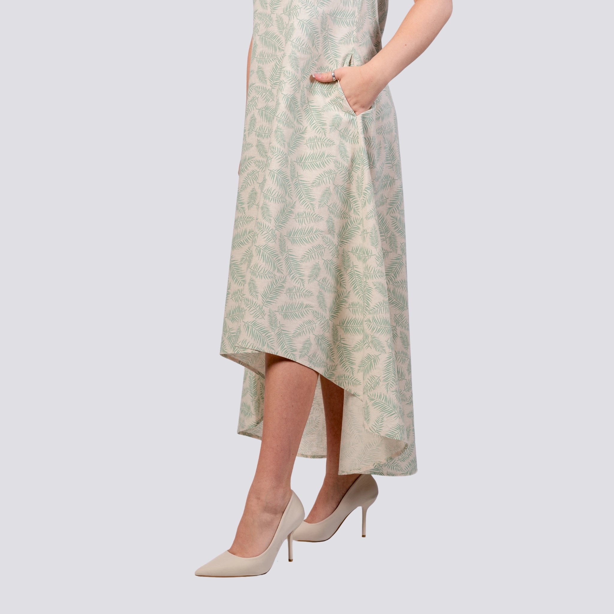 Woman in a Karee Mystic Breeze High Low Linen Cotton Midi Dress, sleeveless U-neck, and beige high heels, mid-section view on a gray background.