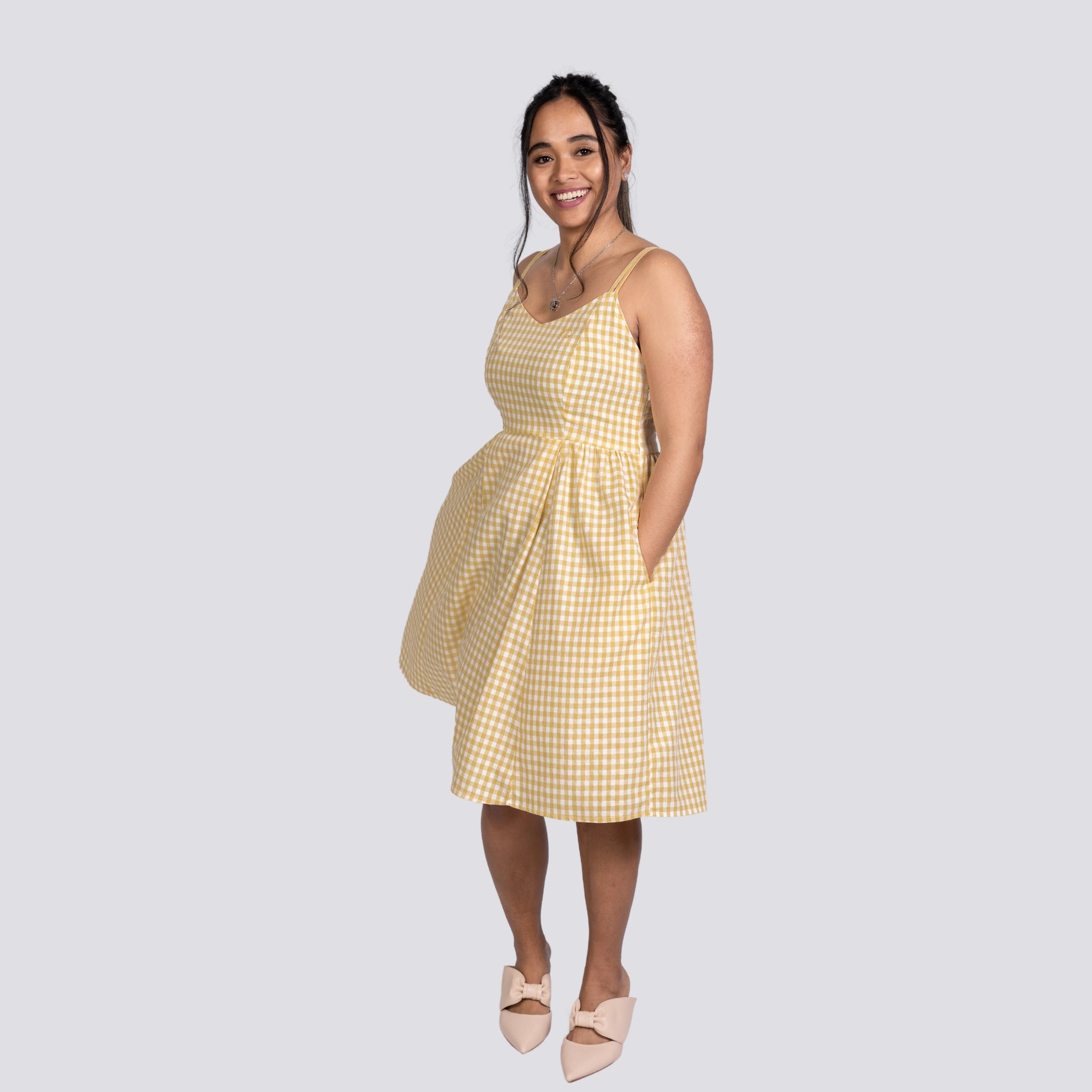 A smiling woman in a Sunshine Chic Yellow Plaid Cotton Mini Dress by Karee and white shoes standing against a gray background.