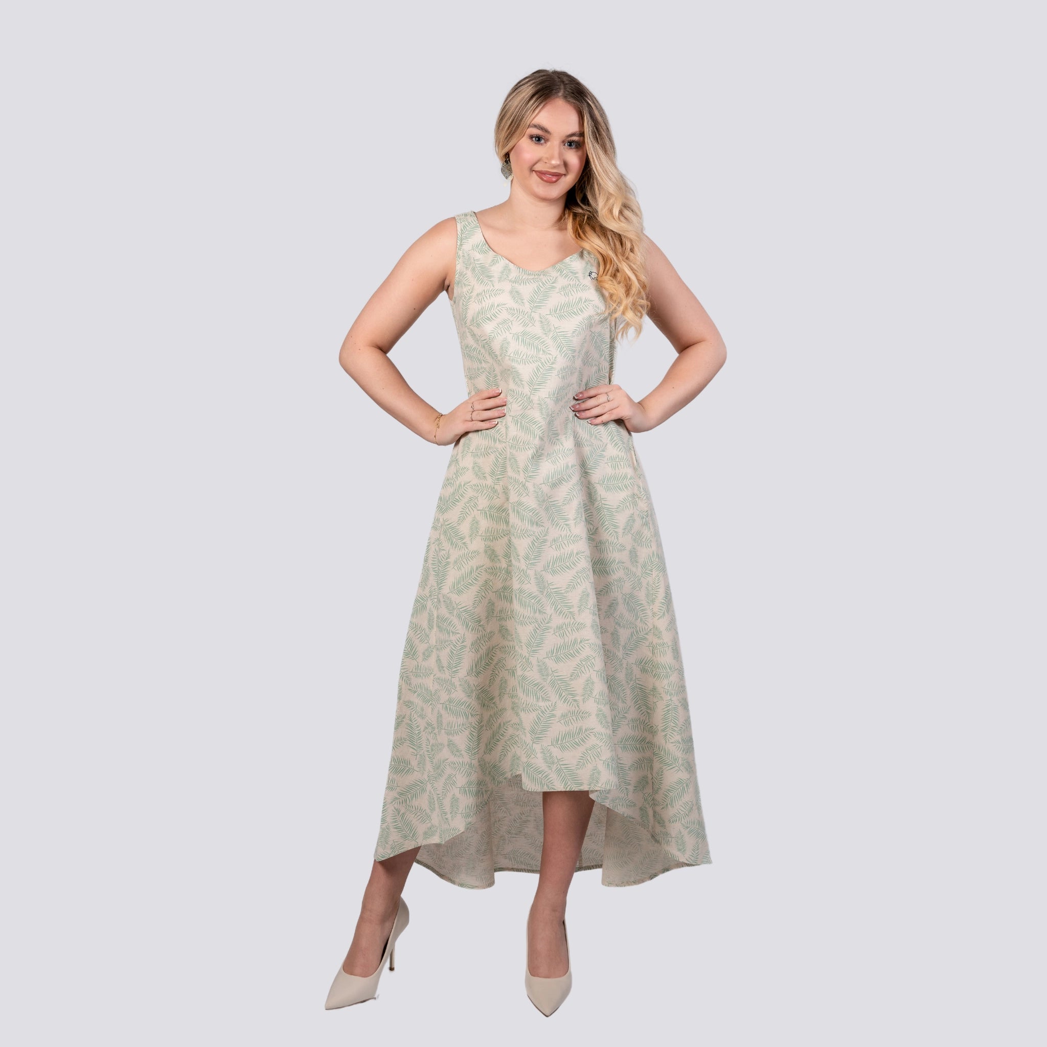 Woman in an elegant sleeveless U-neck leaf-patterned Mystic Breeze High Low Linen Cotton Midi Dress by Karee and white heels, standing with hands on hips, smiling against a light grey background.