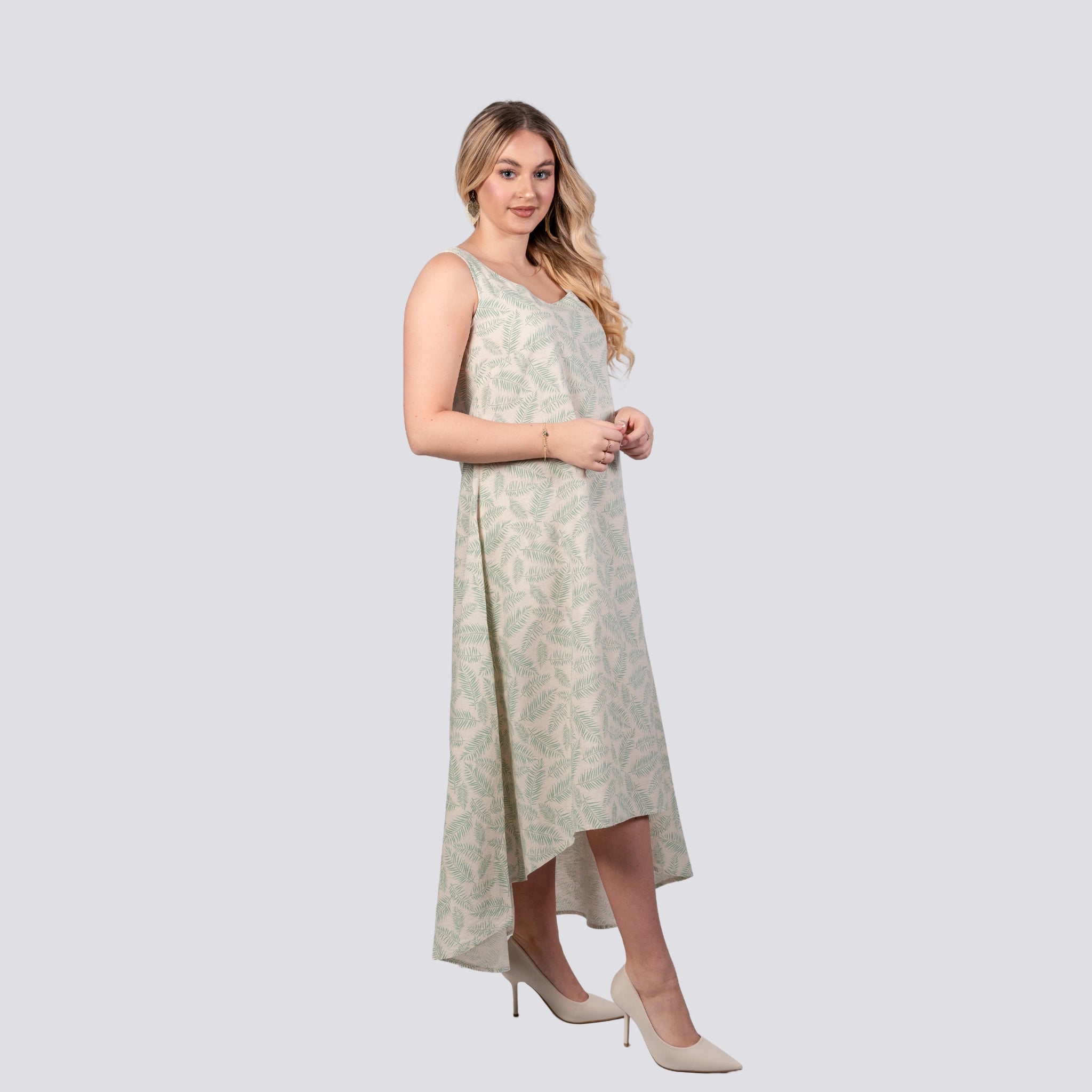 Woman in a sleeveless U-neck Karee Mystic Breeze High Low Linen Cotton Midi Dress and white heels standing against a grey background, looking towards the camera with a slight smile.