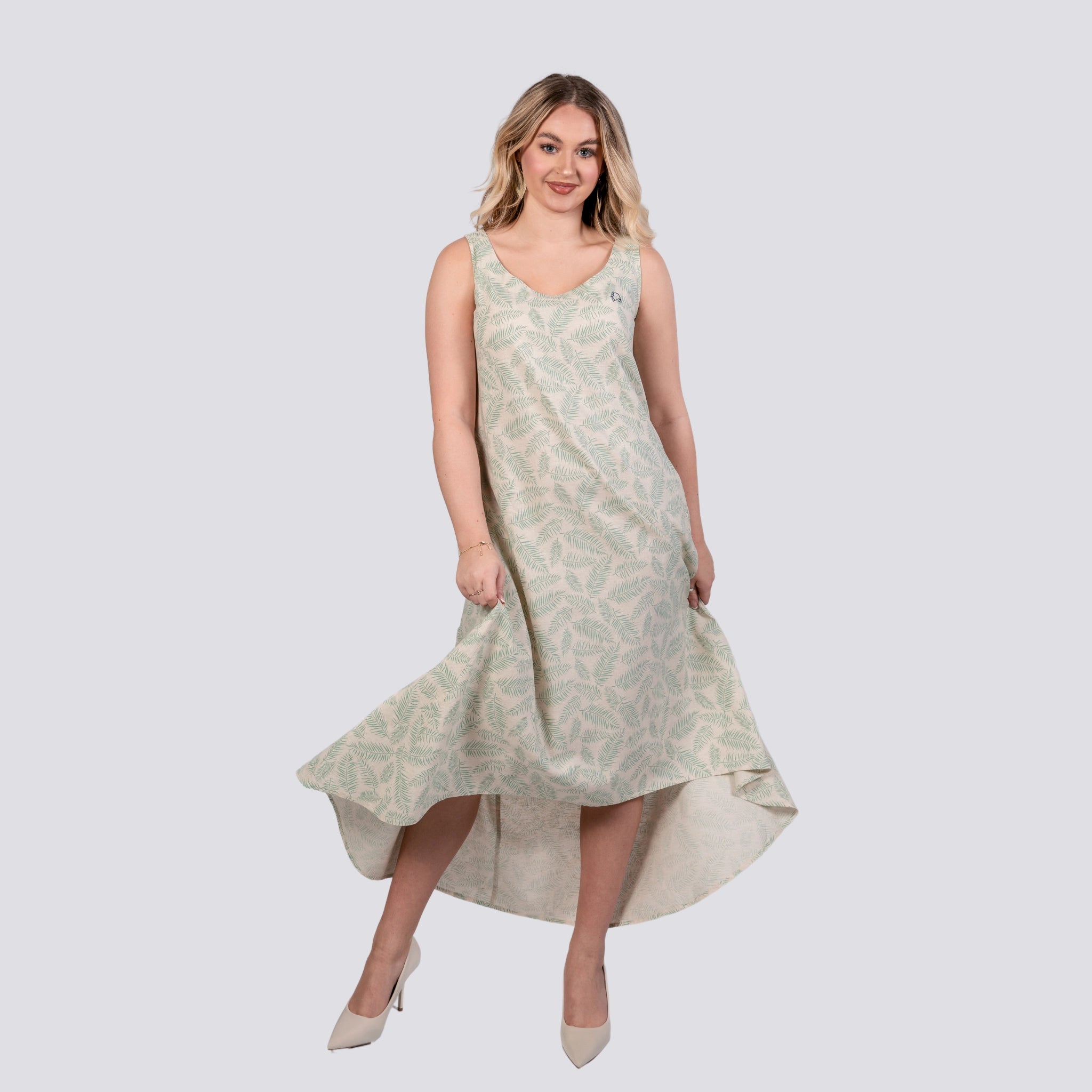 A woman in a flowing Karee Mystic Breeze High Low Linen Cotton Midi Dress, standing against a plain background, smiling and slightly lifting her dress.