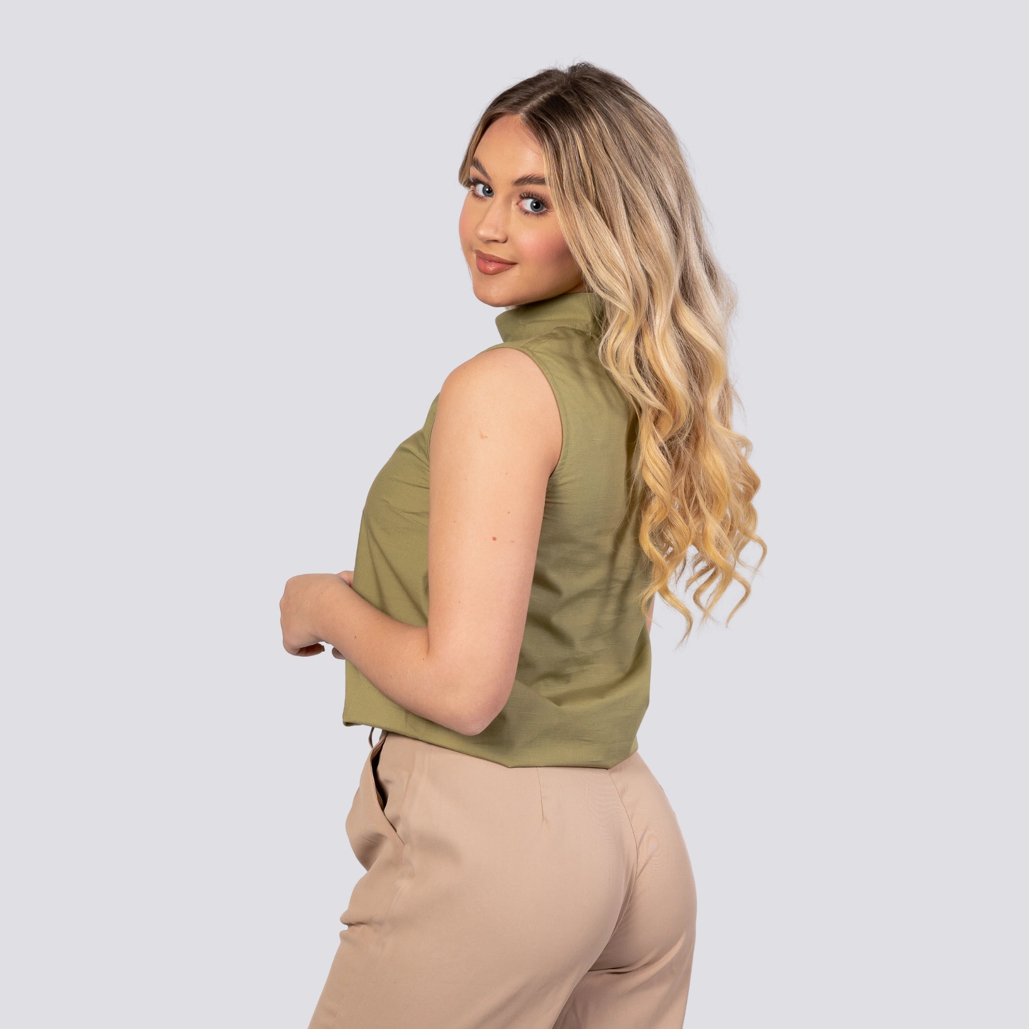 A woman with blonde hair wearing an eco-friendly Karee Pear Green Harmony Sleeveless Top and beige pants, looking over her shoulder with a slight smile, against a light gray background.