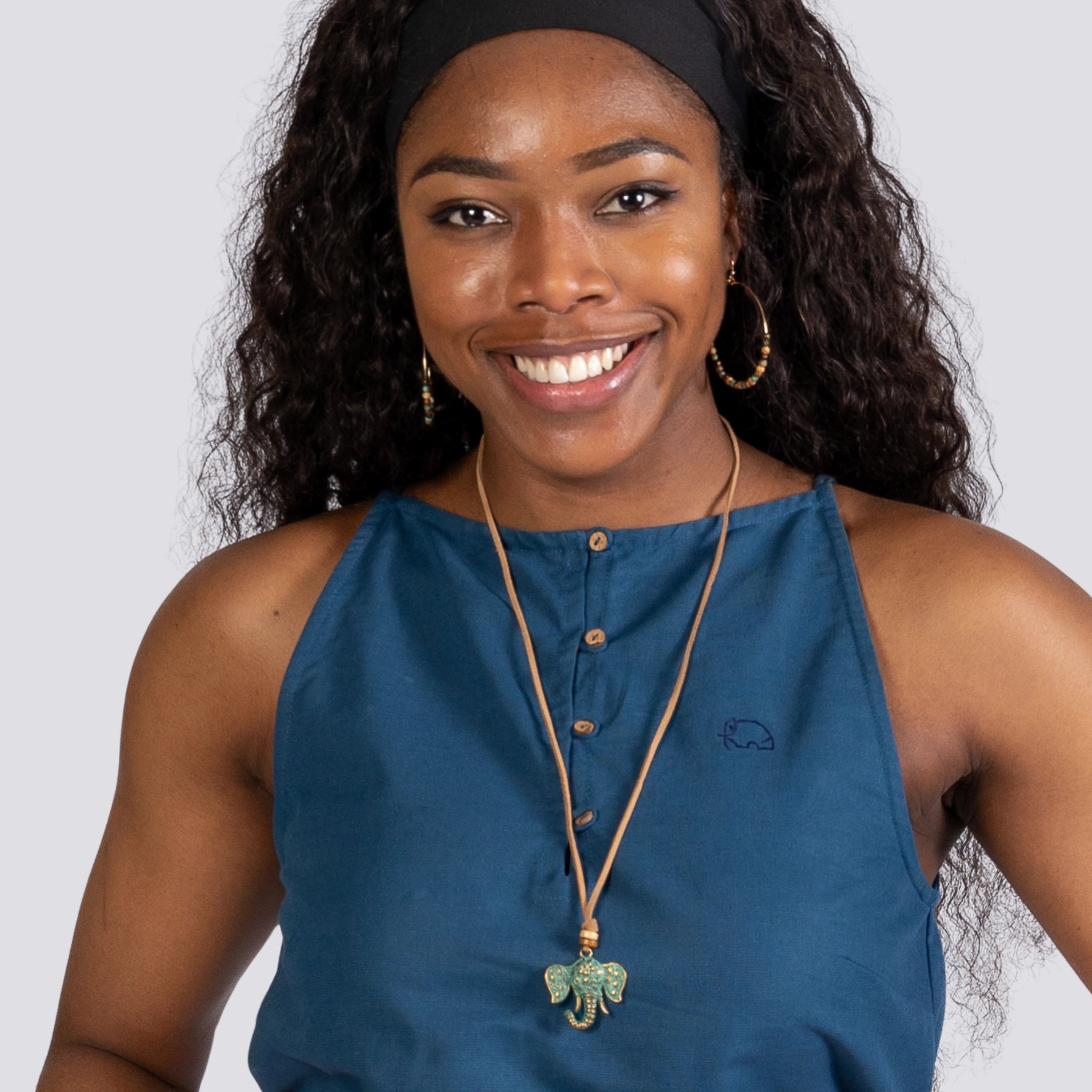 A smiling woman with a headband, wearing a Karee Indigo Twilight Halter Cami Top for Women, hoop earrings, and a necklace with a green pendant.
