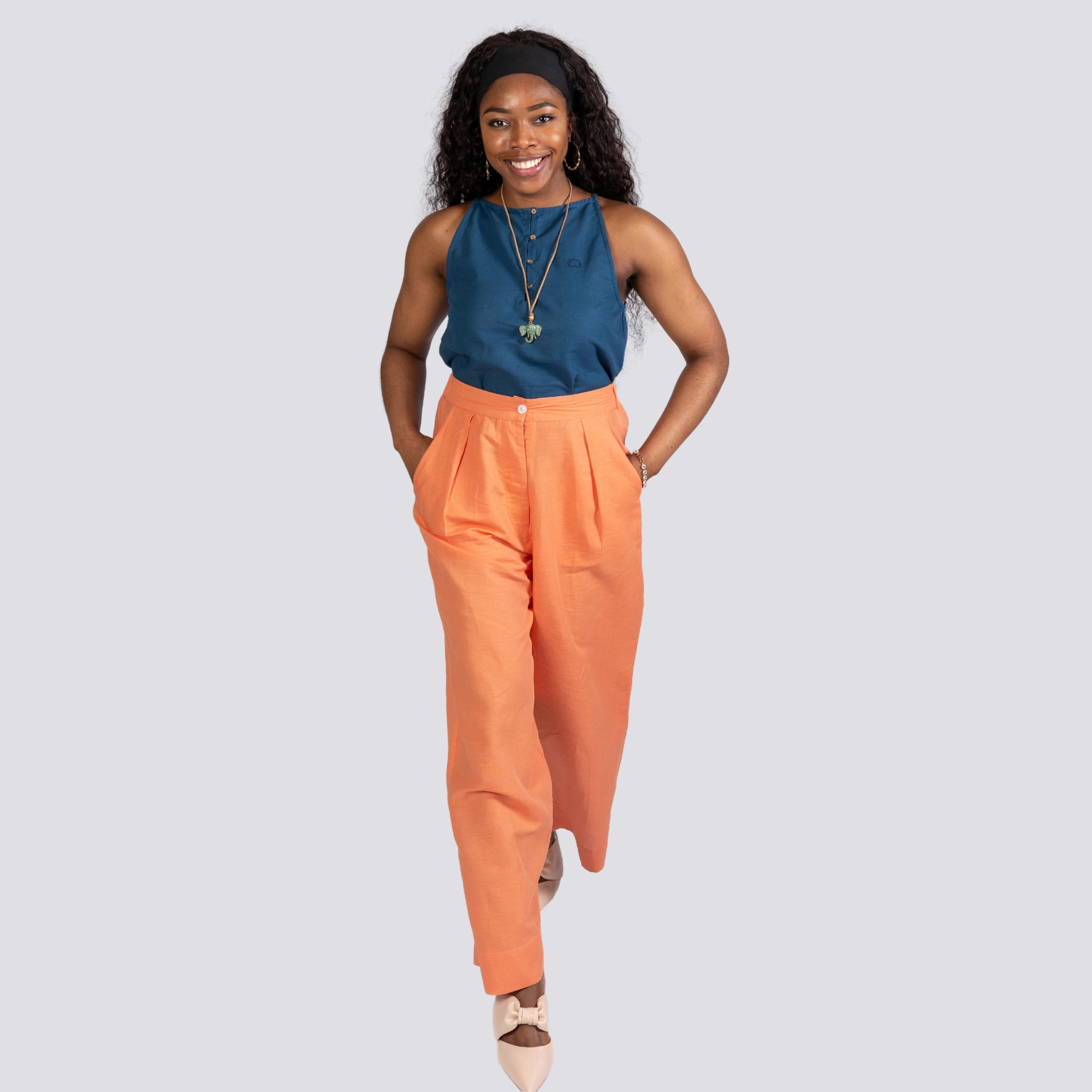 A smiling woman standing confidently, wearing a Karee Indigo Twilight Halter Cami Top For Women, orange trousers, and beige heels, on a grey background.