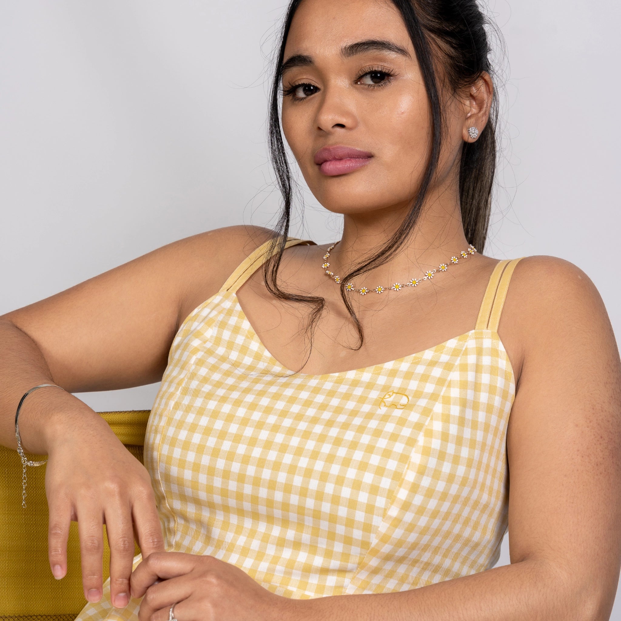 A woman with long dark hair, wearing a limited-edition Karee Sunshine Chic Yellow Gingham Cotton Mini Dress, sits with one arm resting on a chair. She has a neutral expression and is adorned with earrings and a necklace.