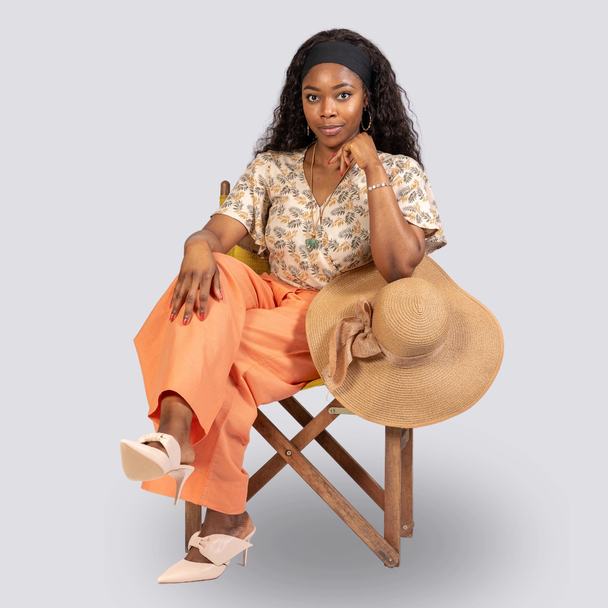 A woman sits on a wooden stool, wearing a ChicPalms Women's Eco-Friendly Wrap Top - Biscuit Bliss by Karee, orange pants, headband, and holding a straw hat, with one shoe off.