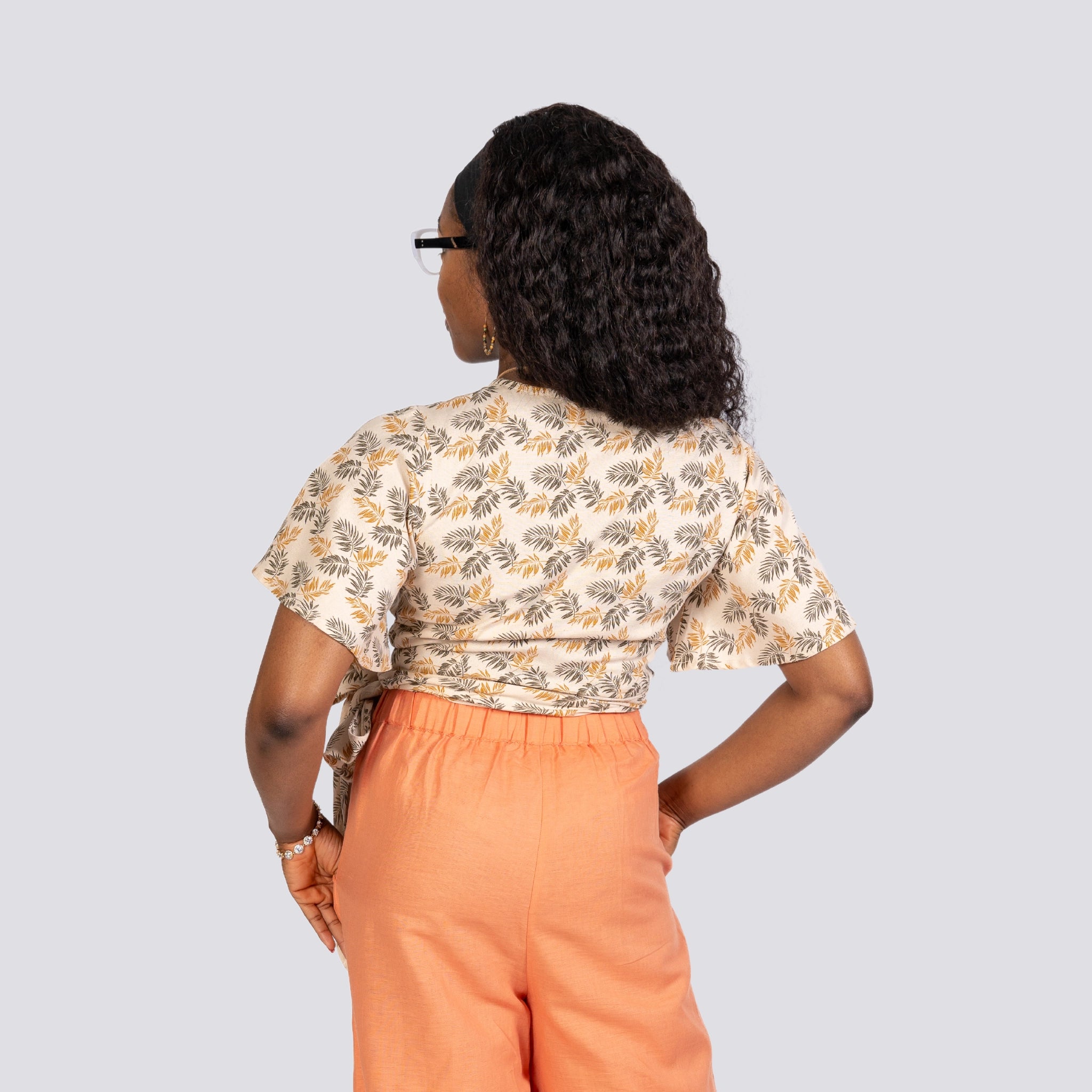 A woman viewed from behind, wearing glasses and dressed in a ChicPalms Women's Eco-Friendly Wrap Top - Biscuit Bliss and orange pants, standing against a gray background.