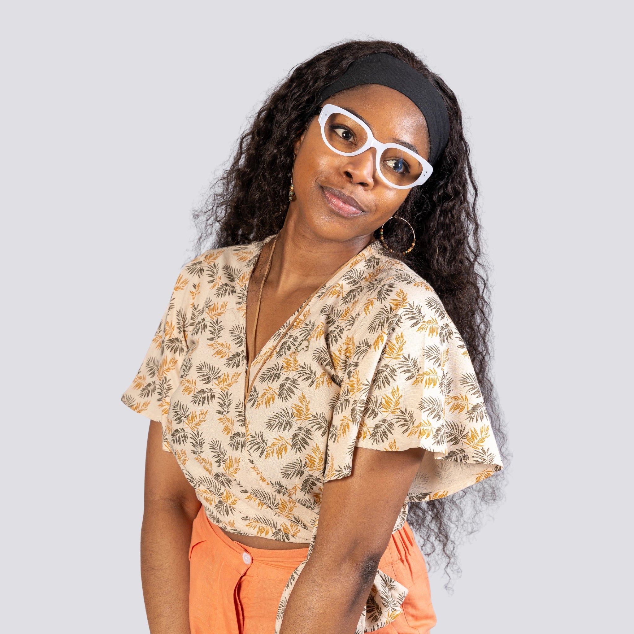 A young woman wearing a ChicPalms Women's Eco-Friendly Wrap Top - Biscuit Bliss by Karee and white sunglasses, paired with an orange skirt, smiles gently against a gray background.
