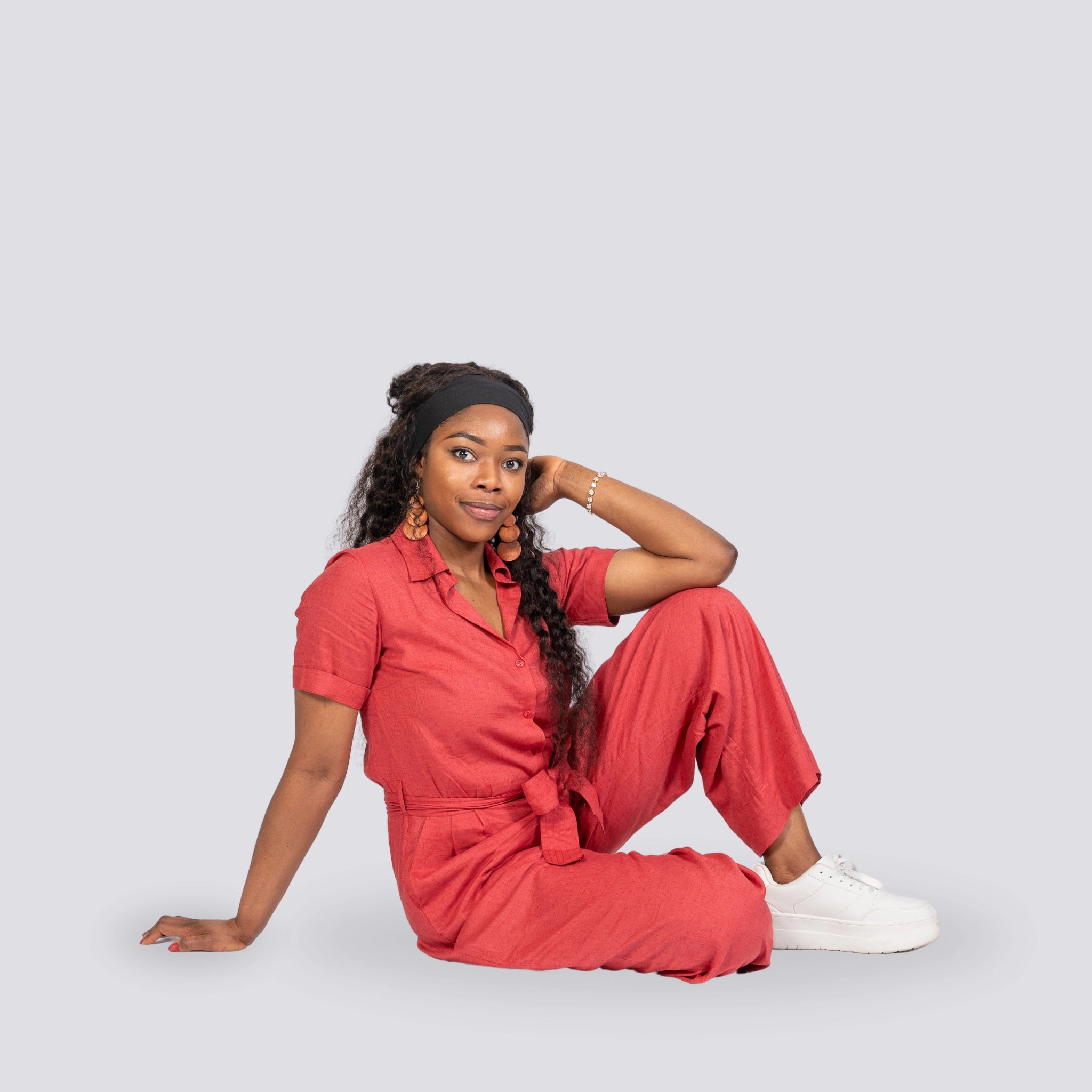 A woman in a Milano Red Serenity Viscose Linen Jumpsuit by Karee sits on the ground, leaning on one hand, wearing white sneakers and a headband.