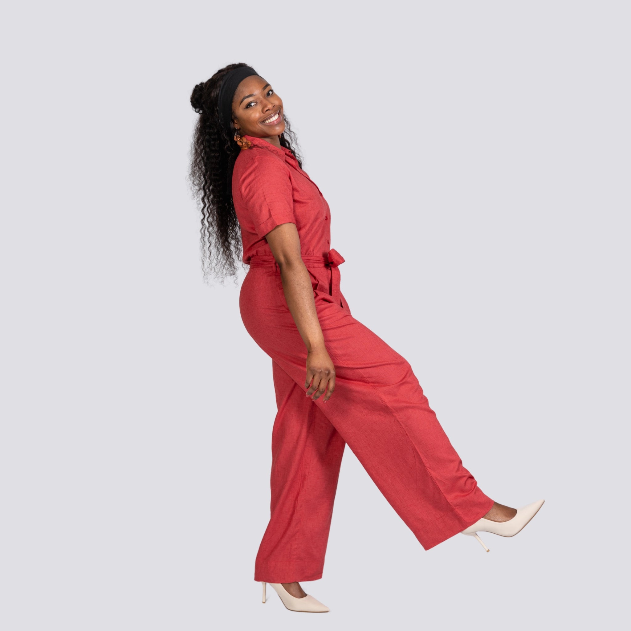 A woman in a Karee Milano Red Serenity Viscose Linen Jumpsuit and white heels smiling and posing playfully against a gray background.
