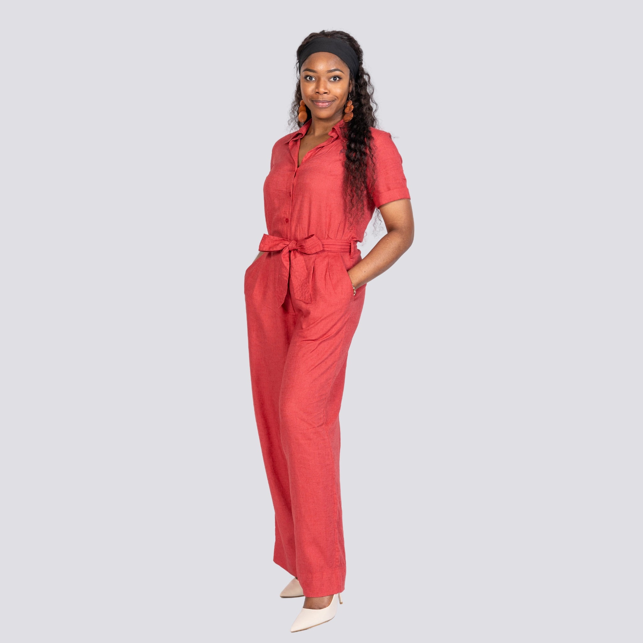 Woman wearing a Karee Milano Red Serenity Viscose Linen Jumpsuit with a tied waist belt, standing confidently with hands on hips against a light gray background.