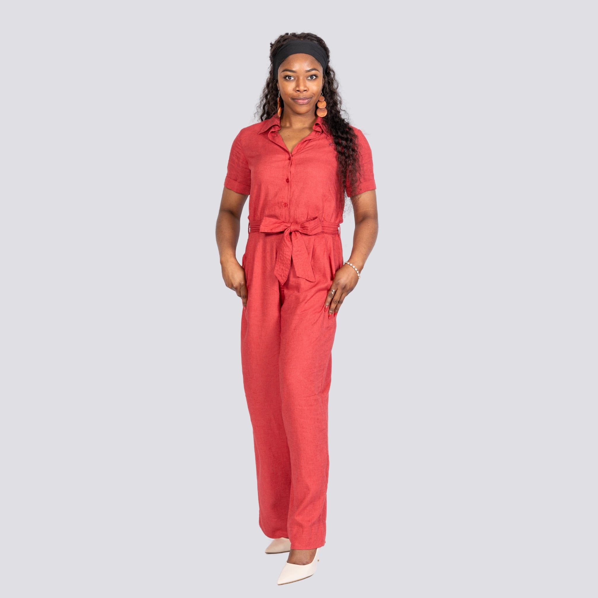 A woman in a Karee Milano Red Serenity Viscose Linen Jumpsuit and white shoes stands against a light gray background, looking at the camera.