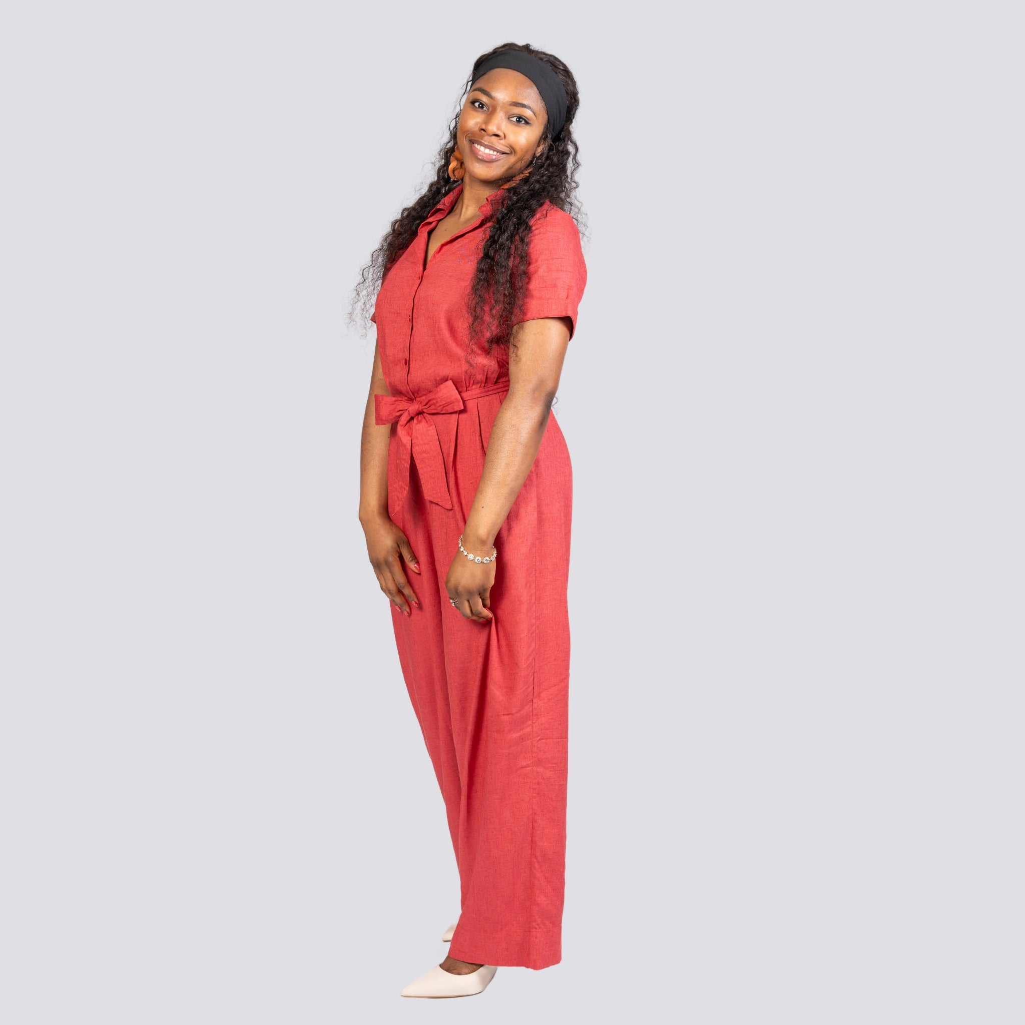A woman in a Milano Red Serenity Viscose Linen Jumpsuit with a tie waist smiling in a studio, standing in a partial profile pose against a light gray background. (Karee)