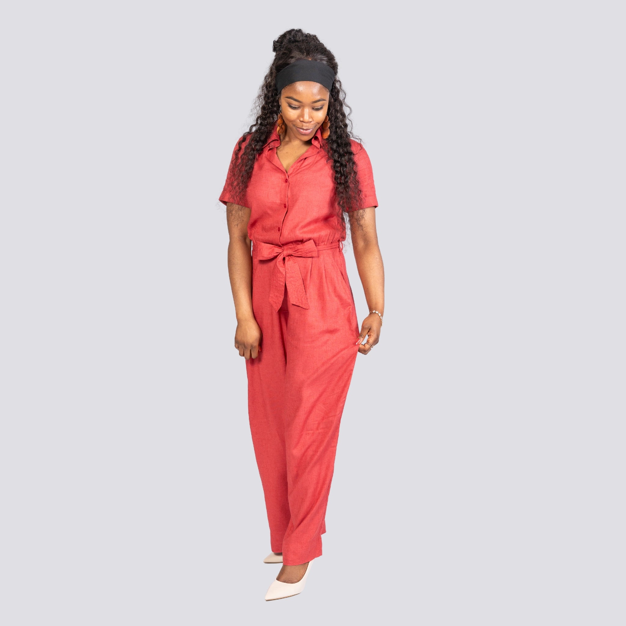 A woman in a Karee Milano Red Serenity Viscose Linen Jumpsuit and white heels looks down while walking, isolated on a gray background.