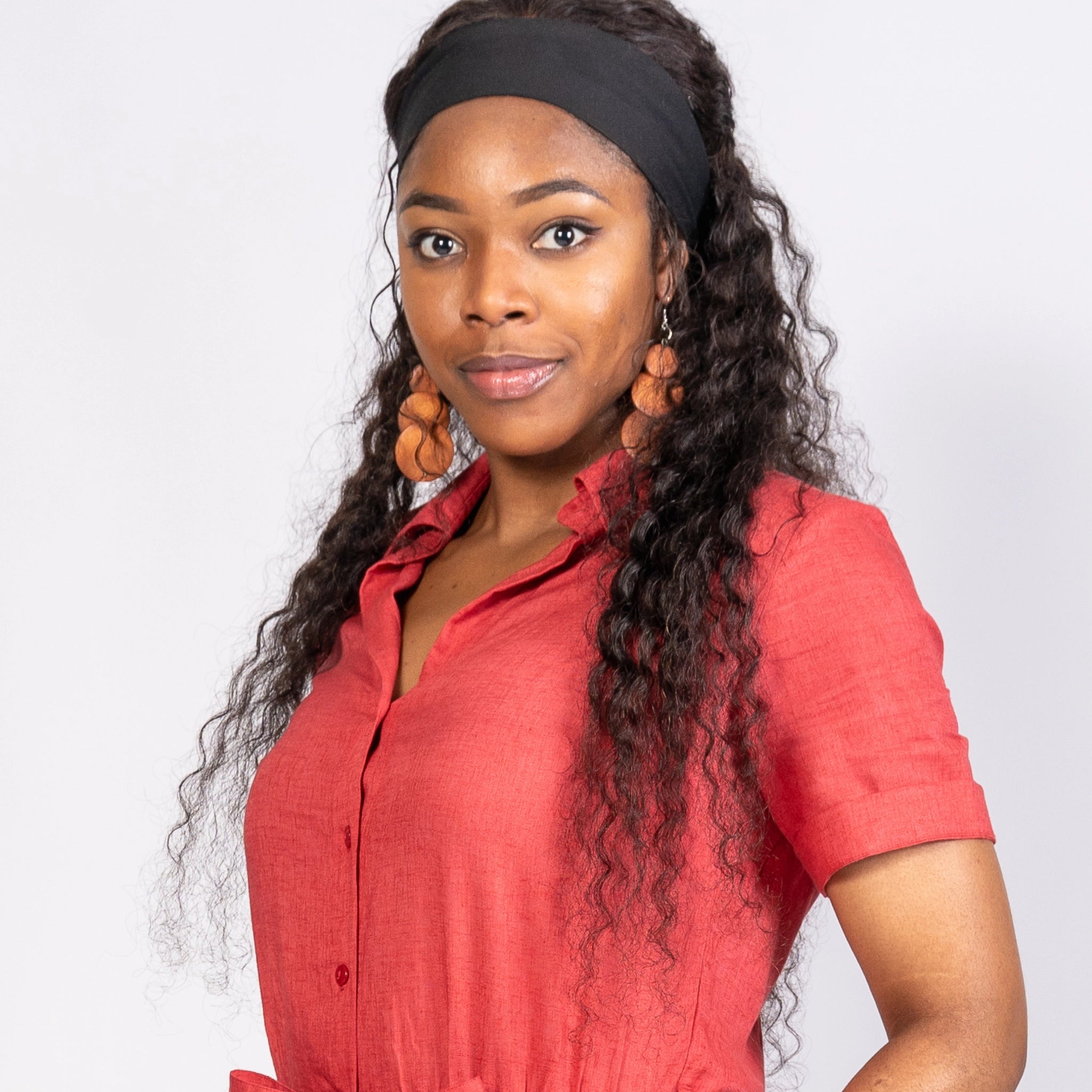 A woman in a Karee Milano Red Serenity Viscose Linen Jumpsuit and black headband, with long curly hair and large circular earrings, stands confidently against a gray background.
